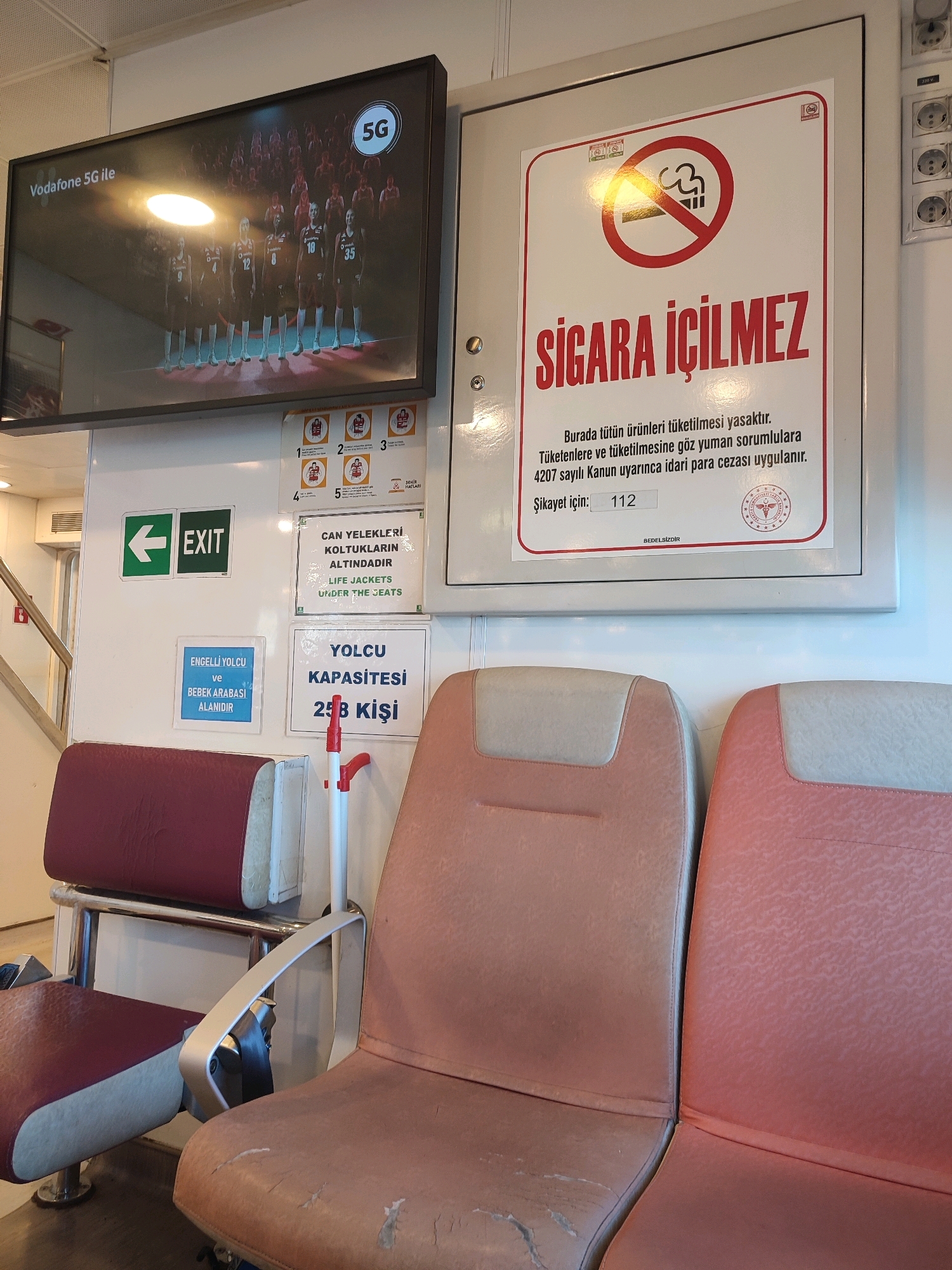 The image depicts the interior of a ferry or public transport vessel. In the foreground, two rows of seating are visible. The seats are upholstered in a red and beige fabric, with some signs of wear and tear. Between the seats, a white pole with a red handle extends upwards, likely part of the seating or a safety feature.
On the wall behind the seats, several signs are displayed. One prominent sign is a "No Smoking" sign with Turkish text. To the left of this, a green sign with the word "EXIT" and an arrow indicates an exit route. Below the "EXIT" sign, a blue sign reads "ENGELİ YOLCU ve BEBEK ARABASI ALANIDIR" (This is a space for passengers with disabilities and for baby strollers). To the right of the blue sign, a white sign states "YOLCU KAPASİTESİ 258 KİŞİ" (Passenger capacity 258 people). Another white sign above it reads "CAN YELEKLERİ KOLTUKLARIN ALTINDADIR" (Life jackets are under the seats).
Above the signs, a large flat-screen television is mounted on the wall. The screen displays a basketball game with players in uniform and the Vodafone 5G logo. The overall environment suggests a public transportation setting, likely on a ferry operating in or around Kadıköy, Turkey, given the Turkish language on the signs. The lighting is artificial and somewhat dim.