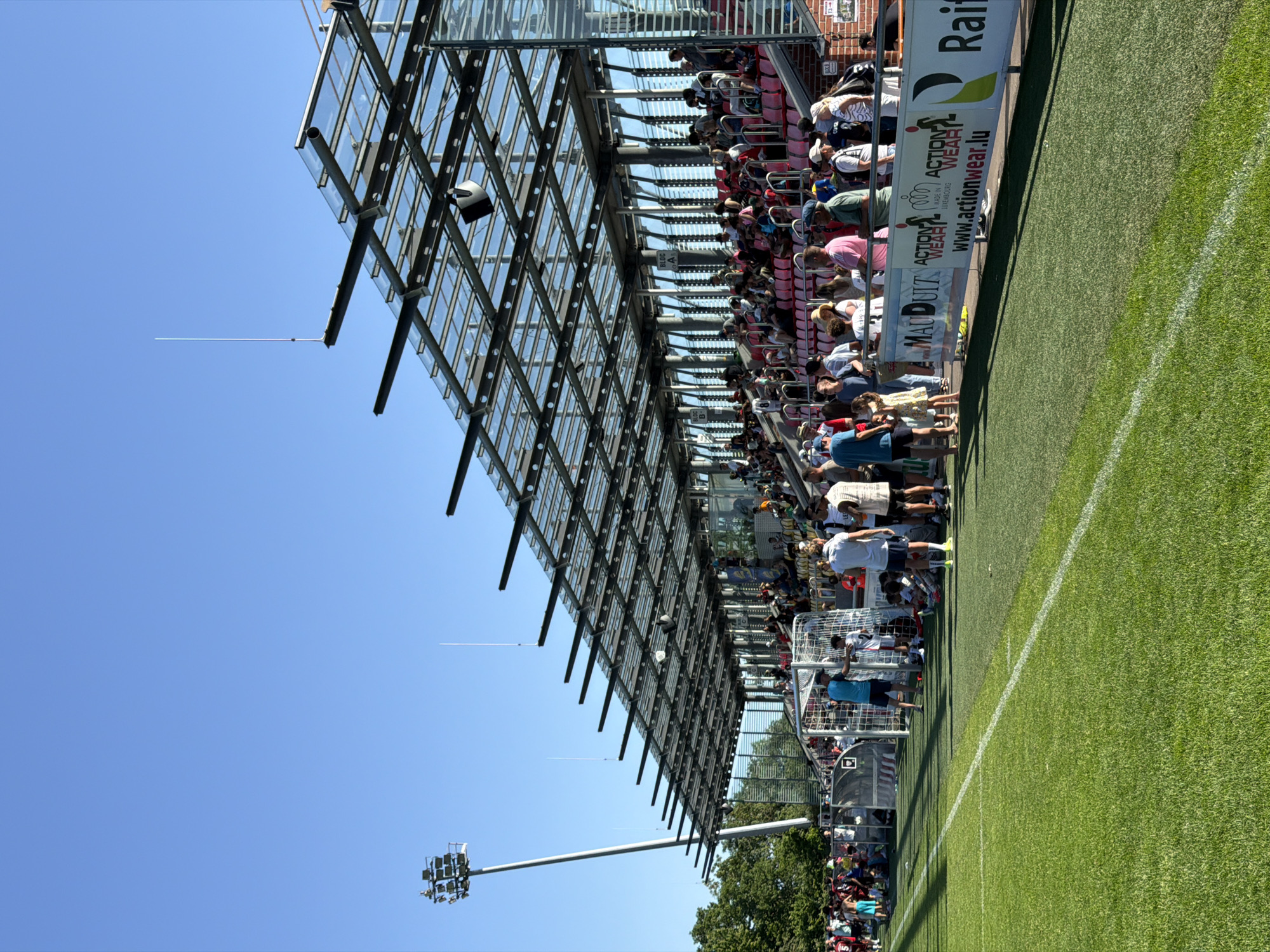 The image depicts a soccer field in Fentange, Luxembourg on a sunny day. Spectators are visible in the covered seating area, a structure with a latticework glass roof. A tall lighting tower stands on the left side of the field, under a clear blue sky. A white sideline is prominent in the foreground. Advertising banners are affixed to the seating area.