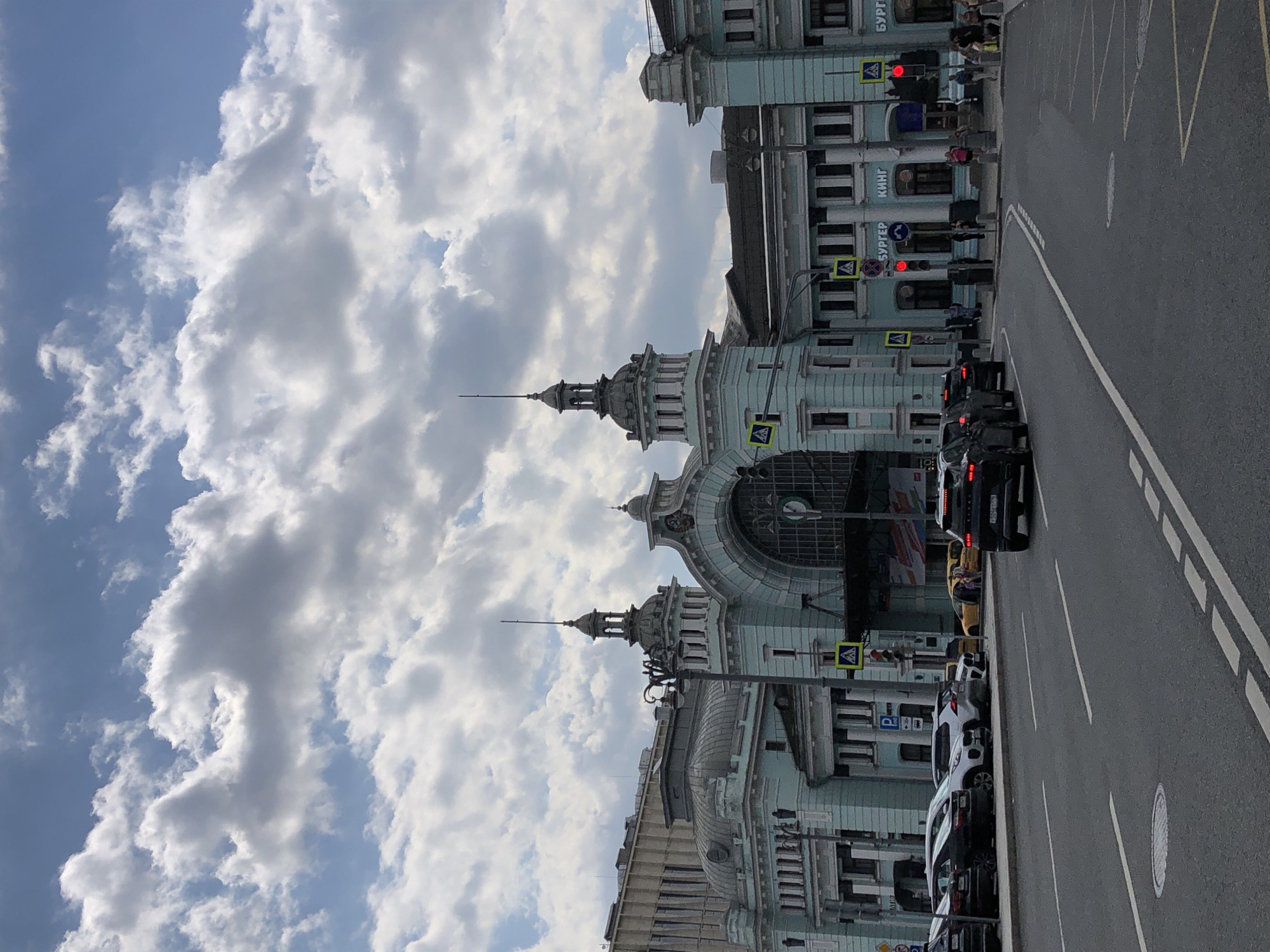 The image depicts a view of the Komsomolskaya Square in Moscow, Russia, featuring the iconic Leningradsky Railway Station. The sky is filled with dramatic, fluffy clouds against a blue backdrop. The facade of the railway station, painted in a pale turquoise hue, is prominent, characterized by its neoclassical architecture with arched windows, decorative spires, and ornate detailing.
In the foreground, a multi-lane asphalt road is visible with white lane markings. Several cars are parked along the side of the road, and a few are in motion. A black SUV is captured in the right lane, and other vehicles, including a white SUV and a yellow taxi, are also present. Pedestrian crossings with yellow cautionary signs are marked on the road. Traffic lights are visible, some showing red.
People are scattered along the sidewalk in front of the station, some walking and others standing. The buildings surrounding the station appear to be modern office blocks or commercial establishments. Signs with Cyrillic lettering, including "БУРГЕР КИНГ", are visible on the lower floors of the station building, indicating commercial businesses. The overall scene captures a busy urban environment with a blend of historical architecture and modern city life.
