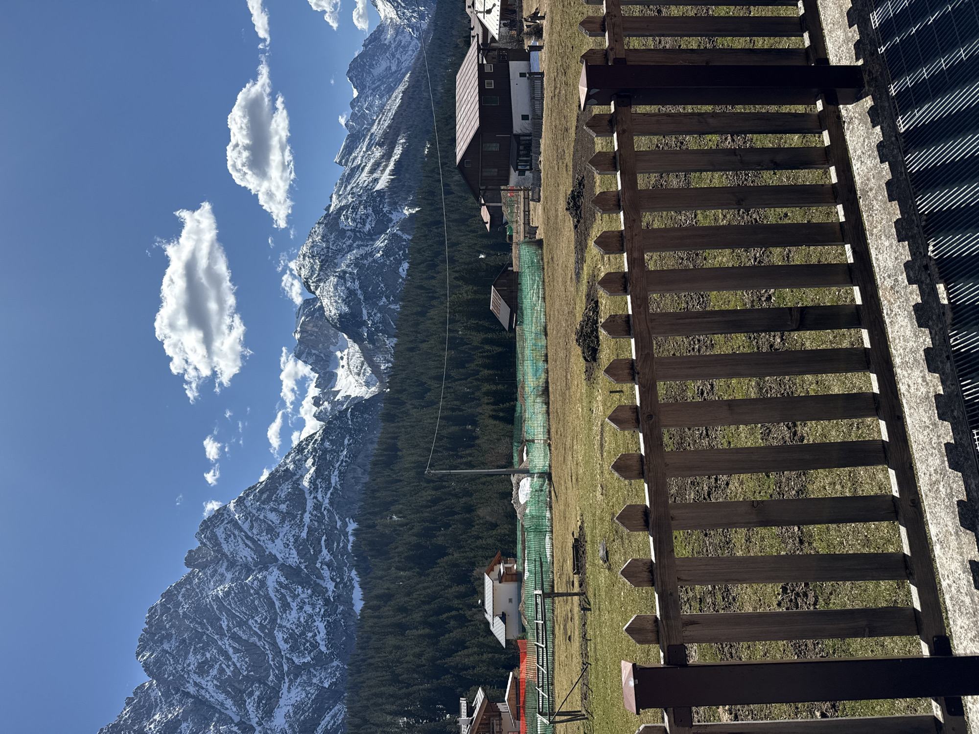The photograph shows a mountain landscape in Sappada, Italy. A wooden fence runs across the foreground. Beyond the fence, a grassy field leads to a dense forest of evergreen trees that ascends the mountainside. The mountains rise sharply in the background, capped with snow and framed by a blue sky dotted with fluffy white clouds. Several buildings are scattered throughout the scene, including what appears to be a traditional Alpine house with a dark roof. A green mesh fence is also visible in the mid-ground.