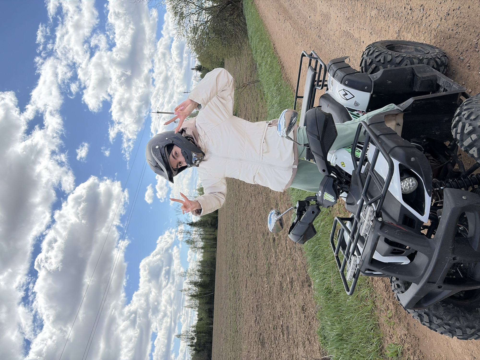 A woman in a helmet and light-colored jacket poses on an ATV in a rural area near Можга, Russia. The ATV is primarily black and white, with front and rear racks. The woman is making a peace sign with both hands. The landscape includes a plowed field, a dirt road, and patches of grass. The sky is blue with puffy white clouds, and power lines are visible.