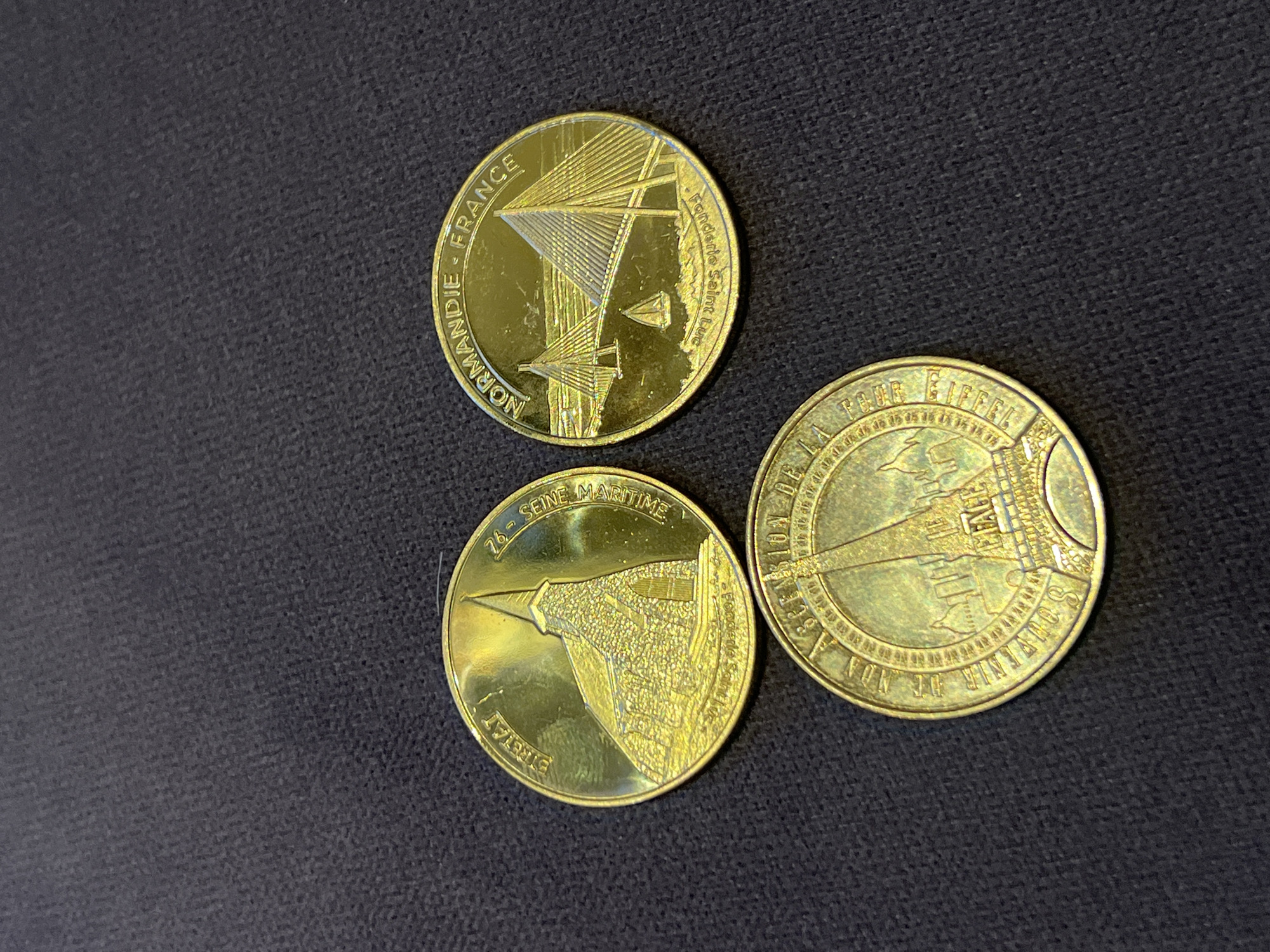 The image shows three golden commemorative coins arranged on a dark, textured surface. The coins are circular and appear to be made of metal with embossed designs.
The coin on the top left depicts a stone church with a steepled roof and arched windows. Text around the rim reads "ETRETAT" and "76 - SEINE MARITIME". Below the church image, partially obscured, is text that seems to read "Fonderie".
The coin on the top right features a modern suspension bridge with multiple cables, likely representing a bridge in Normandy, France. A small sailboat is visible on the water beneath the bridge. The text on this coin reads "NORMANDIE - FRANCE" and "Fonderie Saint-Luc".
The coin on the bottom displays a circular design with the Eiffel Tower prominently featured in the center. Surrounding the Eiffel Tower are smaller structures that appear to be Parisian landmarks. The text around the rim is partially visible and includes "DE LA", "POUR", and "EIFFEL".
There are no people or interactions depicted in the image. The environment is a plain, dark surface, likely a table or fabric. The lighting is warm and highlights the metallic sheen of the coins.
The city of Montfermeil, France is not directly depicted or referenced on any of the coins in this image. The coins appear to be souvenirs from different locations in France.