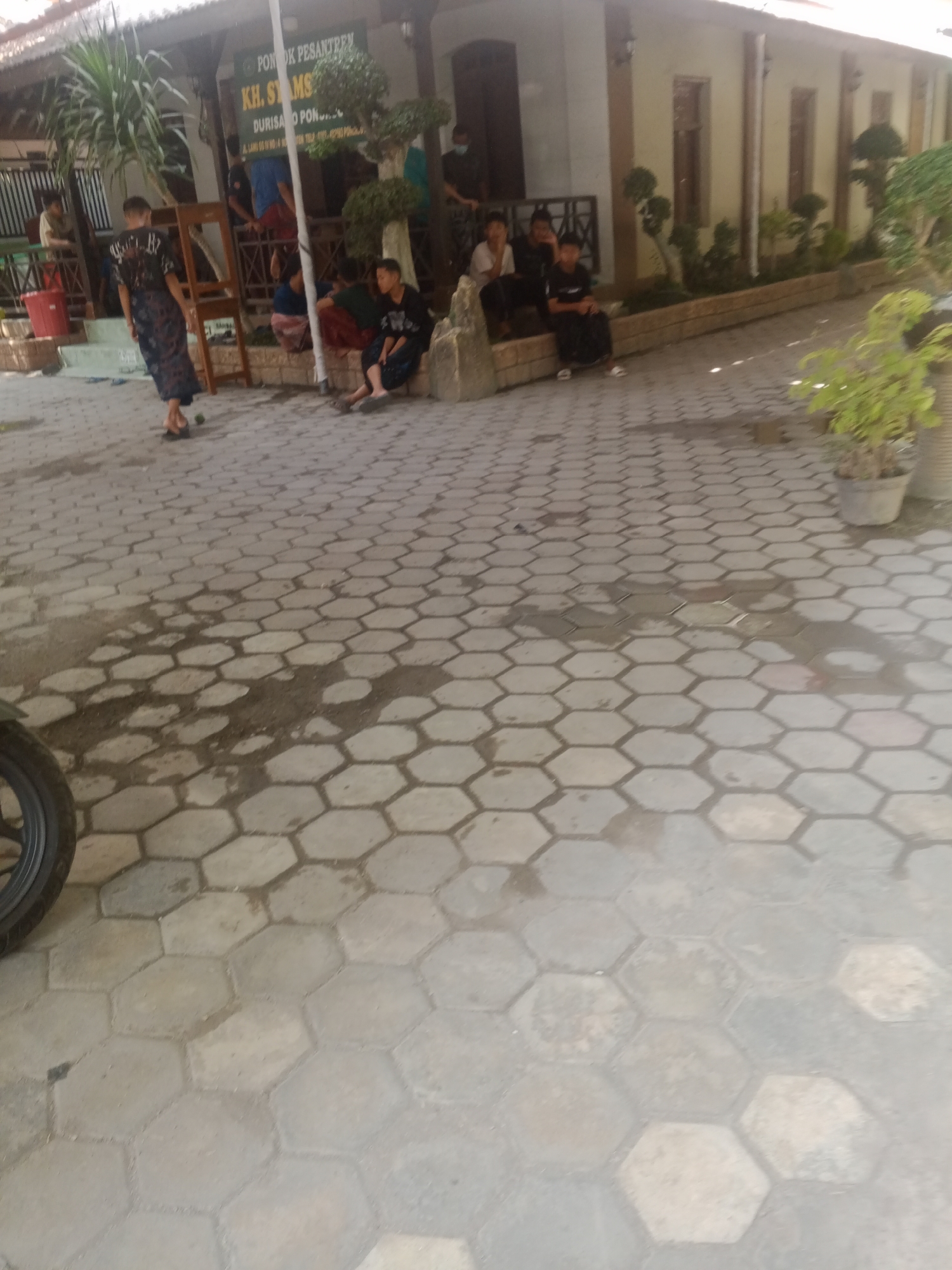 In Banyudono, Indonesia, a street scene unfolds with a group of young men gathered in front of a building identified by a sign as "Pondok Pesantren KH. SYAMS". The building appears to be a pesantren, or Islamic boarding school. Some of the young men are seated on a low wall or steps, while others are standing or walking. One individual, wearing a dark shirt and patterned pants, is walking away from the camera, his back turned. Another young man in a black shirt and pants is sitting with his legs crossed, looking towards the building. The ground is paved with hexagonal tiles, some of which appear wet. A red bucket is visible near a building entrance. The overall atmosphere is casual, with the young men seemingly engaged in a break or social gathering.