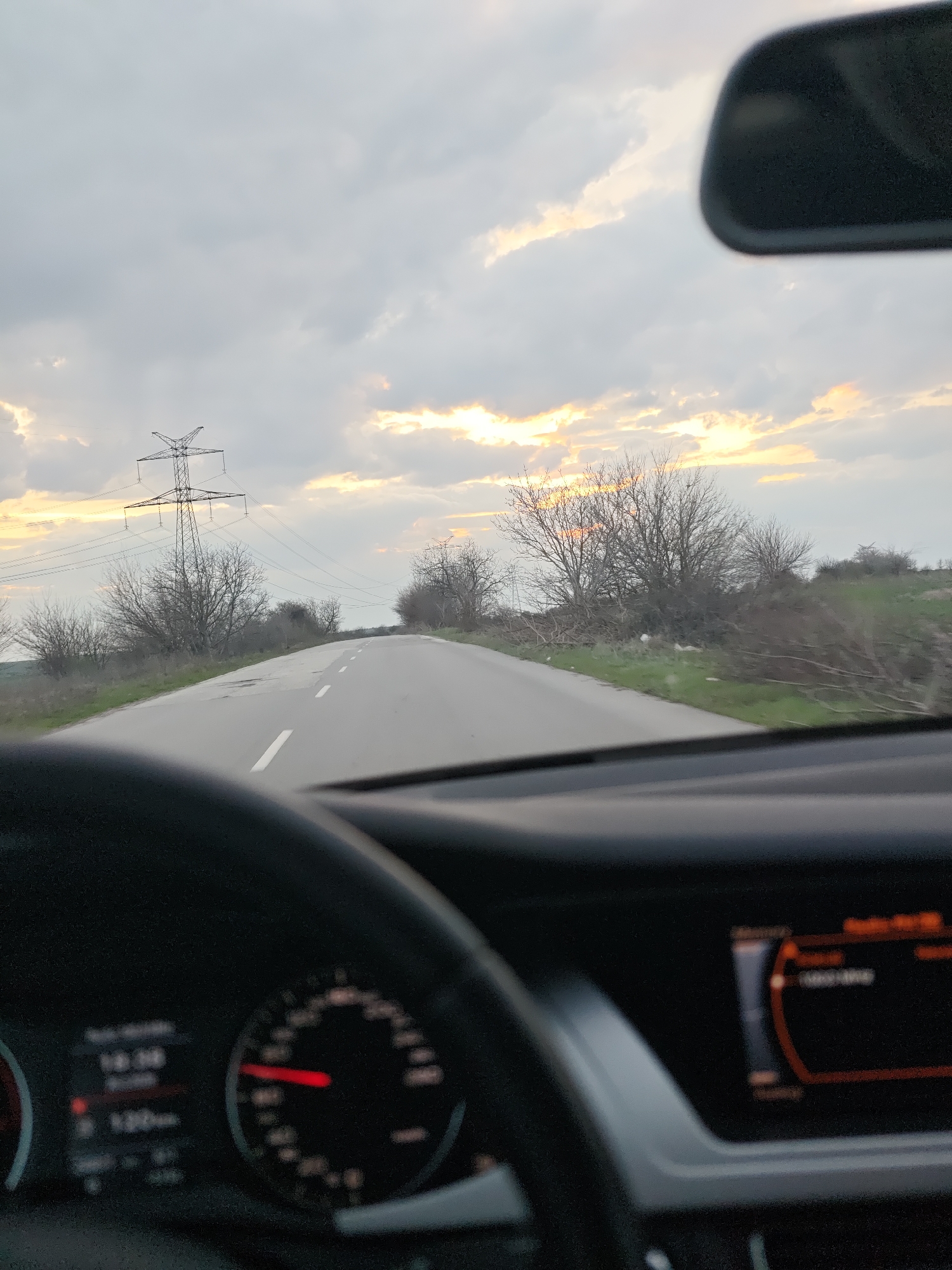 The image captures a forward perspective from inside a vehicle on a paved road located in Gorni Tsibar, Bulgaria. The time is approximately 18:00, indicated by a blurred digital display within the instrument cluster visible behind a dark steering wheel in the lower left. A red needle on the speedometer is partially discernible. A blurred infotainment screen is present on the lower right dashboard, featuring an orange-toned interface. A portion of the vehicle's rearview mirror is visible in the upper right.
The road is asphalt, marked with white dashed center lines, and extends into the distance, exhibiting a slight upward grade and gentle curve to the right. Both road shoulders are covered in green vegetation, with numerous bare deciduous trees and shrubs lining the route. On the left side of the road, a prominent metal electricity transmission tower stands, supporting multiple power lines that traverse the landscape. The sky is predominantly cloudy, with distinct areas of orange and yellow light on the horizon, consistent with twilight conditions.