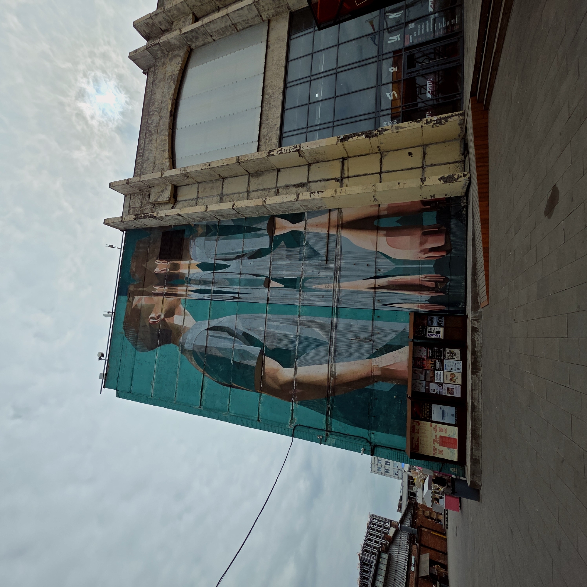 The image shows a building in Saint Petersburg, Russia. The building has a large mural of a man and woman on its side. There is a small display of posters and flyers to the right of the mural. The sky is cloudy and overcast. The building is old and weathered, with peeling paint and cracks in the concrete. The mural is painted in shades of blue, green, and brown. The man in the mural is wearing a hat and a shirt. The woman is wearing a dress. The posters and flyers are advertising various events and businesses. The building is located on a paved street. Other buildings are visible in the background.