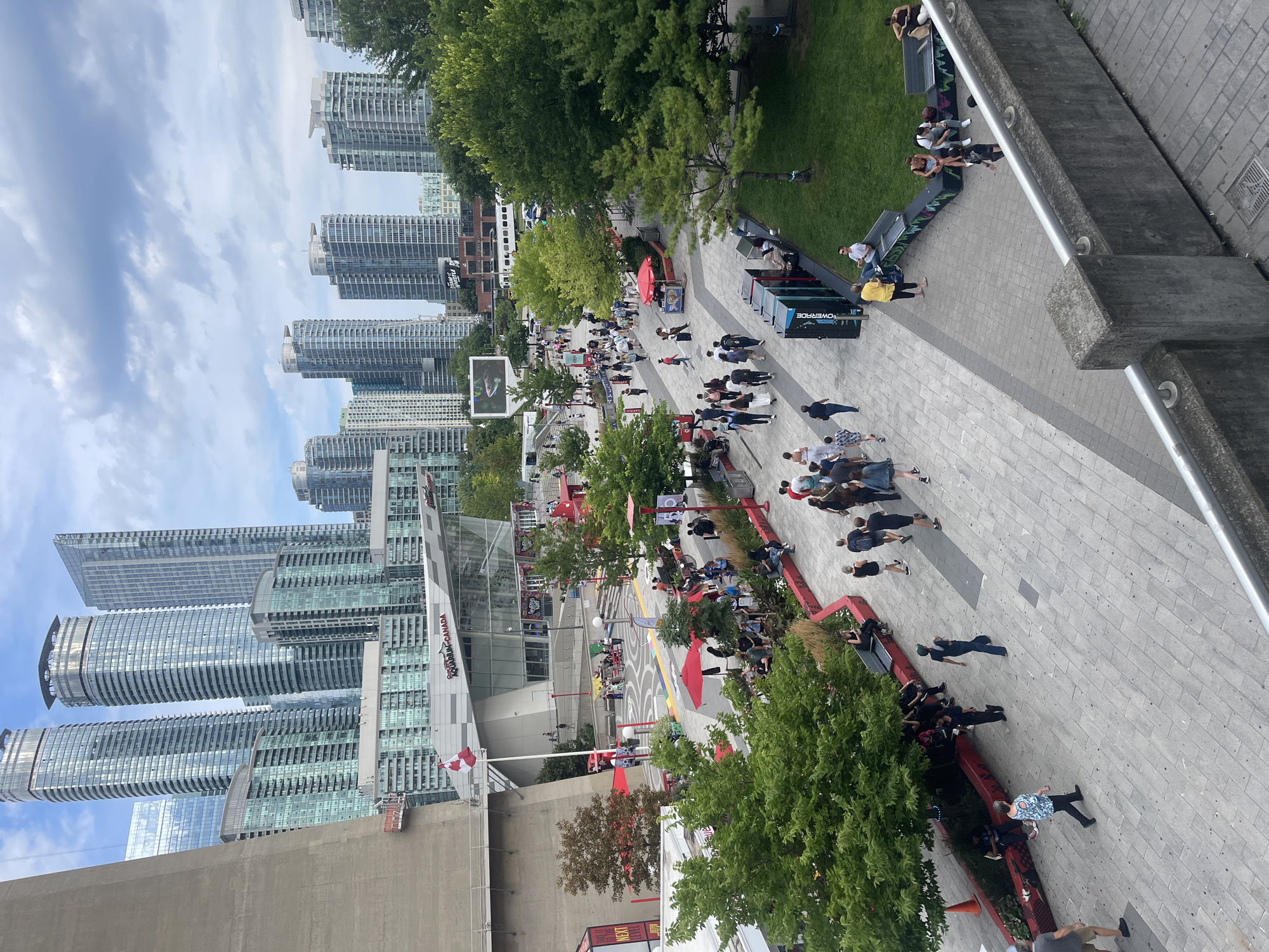 This image depicts a bustling pedestrian walkway in Toronto, Canada, likely near the CN Tower. In the background, modern skyscrapers pierce the cloudy sky. A Canadian flag flies proudly in front of a building labeled "Ripley's Aquarium of Canada".
The foreground is a wide, paved path lined with trees and outdoor seating areas. Numerous people are walking, sitting, and socializing, dressed in casual attire. Some individuals are gathered around red umbrellas, suggesting vendor stalls or shaded seating. The overall scene conveys a vibrant urban atmosphere with a mix of tourists and locals enjoying the outdoor space.