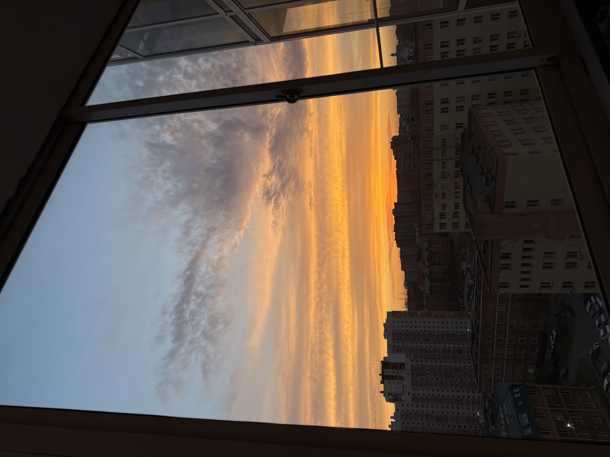 From an elevated position, possibly inside an apartment in Krasnoyarsk, Russia, the view through an open window showcases a vivid sunset scene. The skyline is dominated by apartment buildings, varying in color from light yellow to orange-brown, and some are brick red. Between the window frames, the sunset paints the sky with bands of orange, yellow, and greyish clouds. The city's layout includes streets with parked vehicles interspersed among the buildings. The photograph is taken at an angle, with the window forming the foreground.