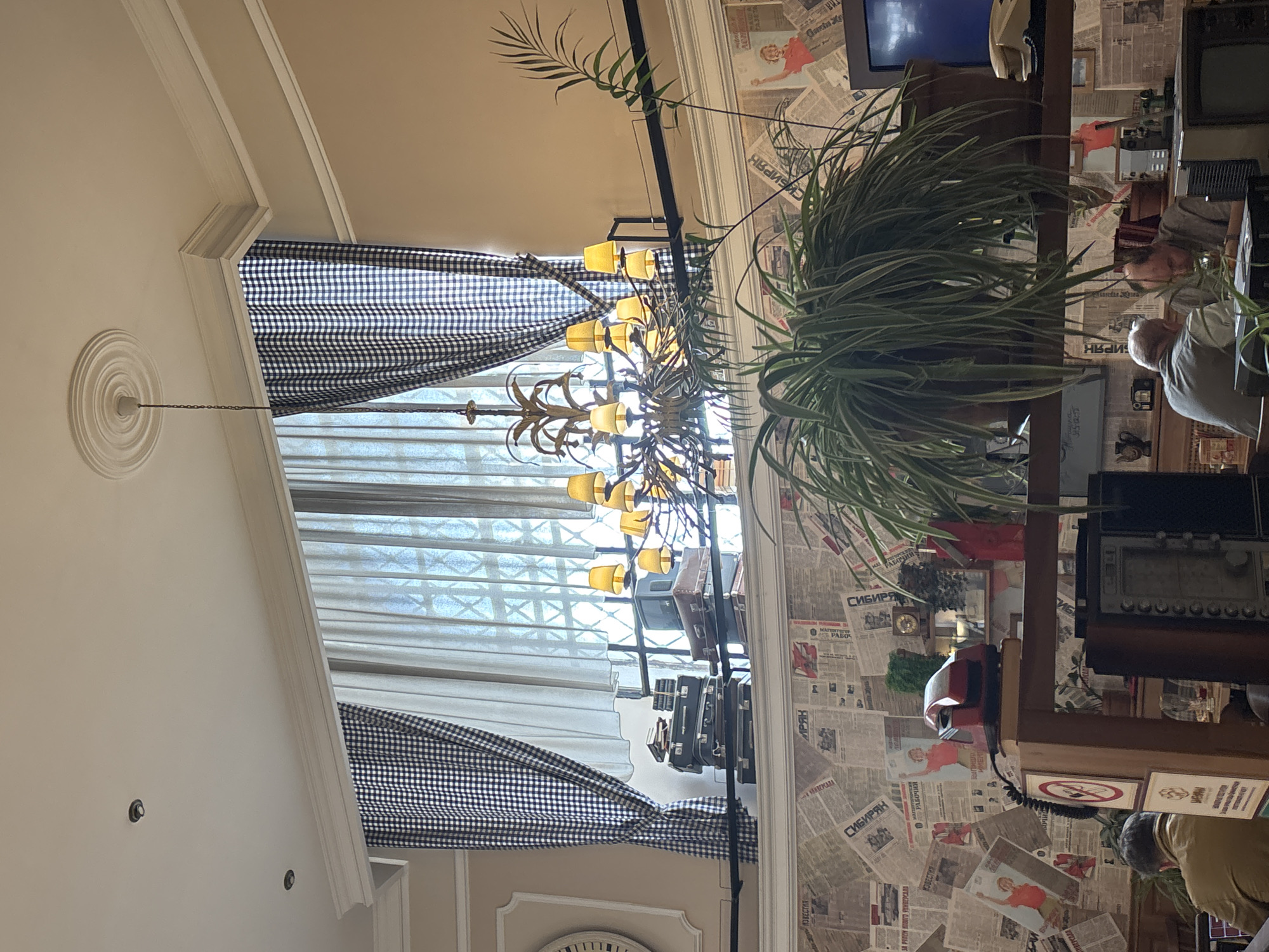 The image shows the interior of a restaurant or cafe in Moscow, Russia. The room is decorated in a vintage style, with walls covered in newspaper clippings. A chandelier hangs from the ceiling, and a large plant sits on a shelf. A television and various other retro items like radios and vintage suitcases are displayed on the shelf. Curtains are hanging along the windows. Patrons can be seen at the side of the image.