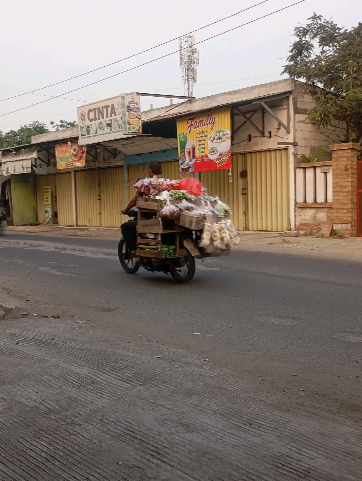 A man is riding a motorcycle laden with goods on a paved road. The motorcycle has a wooden cart attached to the rear, overflowing with various items, including what appears to be produce in plastic bags and bundles of dried goods. The man is wearing dark clothing and is positioned on the seat of the motorcycle, facing away from the camera. In the background, there are buildings with yellow roll-up doors and signage, including one that reads "CINTA." A tall cell tower is visible in the distance, and trees line the side of the road. The sky is overcast. This scene is typical of local commerce in the city of Babelan, Indonesia, where street vendors utilize motorcycles to transport and sell goods.