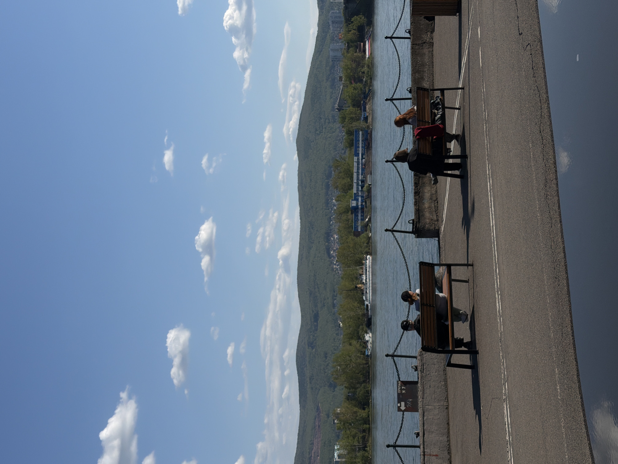The image captures a scene along the Yenisey River embankment in Krasnoyarsk, Russia. Two sets of people are sitting on benches, separated by a distance, along a concrete walkway secured by a chain railing. The first bench has two people, one with long red hair and a person wearing a red garment. The second bench also has two people sitting on it, possibly viewing the river. Behind the walkway, the Yenisey River flows, with a green, mountainous landscape in the background, extending into the distance. A clear blue sky with scattered clouds is visible above. The picture has been taken in a vertical orientation.