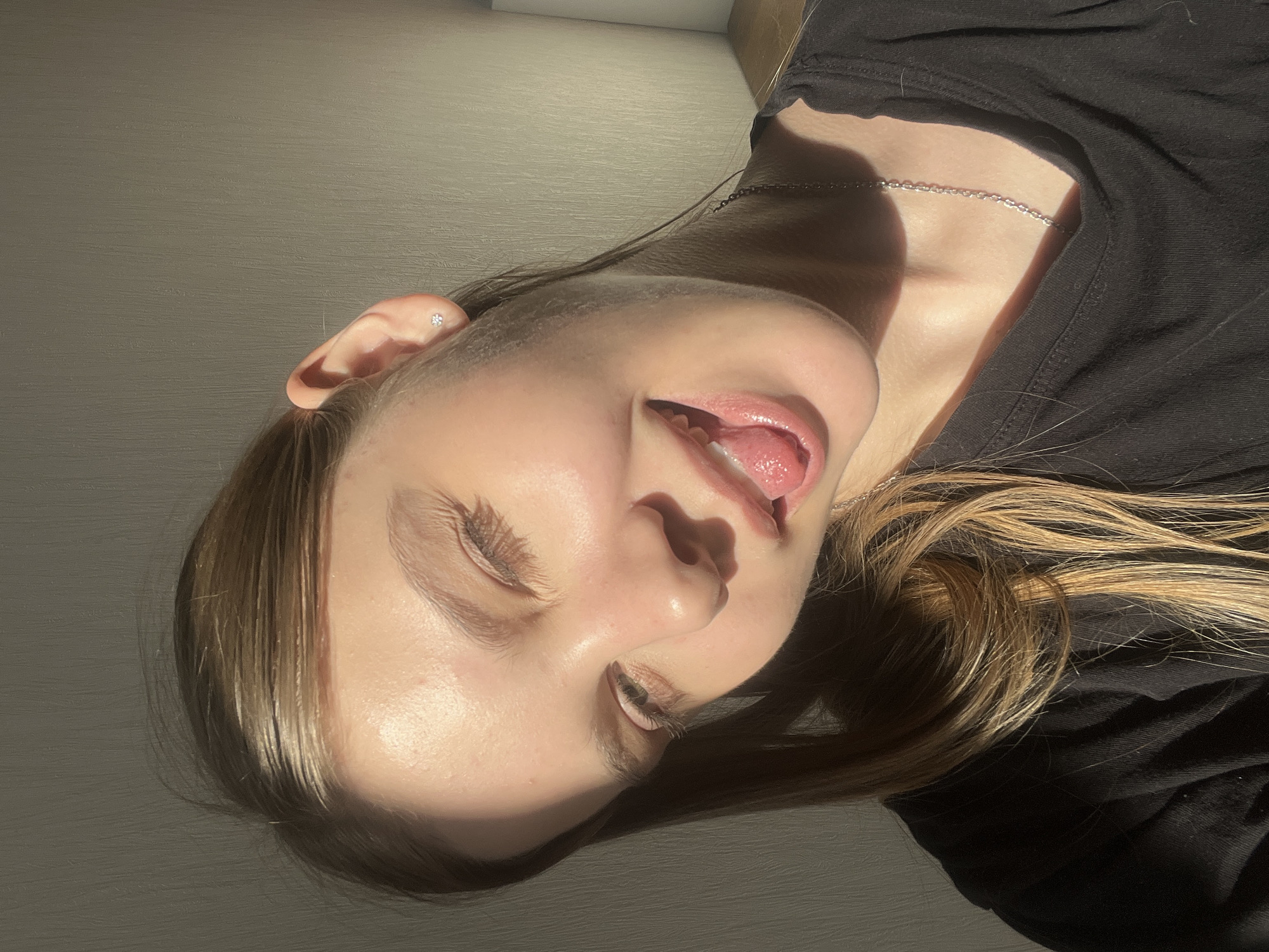 A young, fair-skinned person is positioned at an angle in the frame, facing slightly downward and to the right. Their eyes are mostly closed, with lashes visible, and their mouth is open, revealing their tongue. They have a small earring in their left ear. The person is wearing a black shirt, and a silver chain necklace is visible around their neck. Light brown and blonde hair is partially visible. The background is a light gray or beige wall. The lighting is such that there are distinct areas of light and shadow on the person's face and clothing. The location in the city of Votkinsk, Russia is unknown.