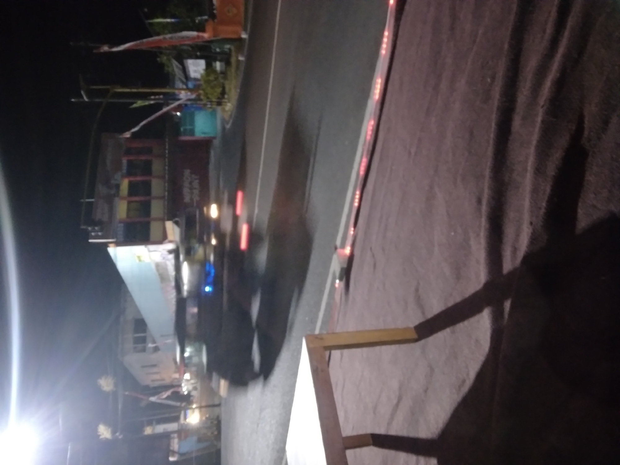 This is a nighttime, low-light, motion-blurred photograph taken in Pacet, Indonesia. The foreground features a light-colored wooden table with dark shadows cast across a dark, textured surface, likely a rug or cloth. In the mid-ground, a road is visible with a moving vehicle, its lights blurred into streaks of white and red. The vehicle appears to be a car or van. Behind the road, a building with multiple windows and a light blue facade is prominent, with the text "NIPPON PAINT" visible on its side. Flags are flying from poles in front of the building. Streetlights and the bright glare of a light source are also present, contributing to the overall low-light conditions and blurring. The environment suggests an urban or semi-urban setting at night.