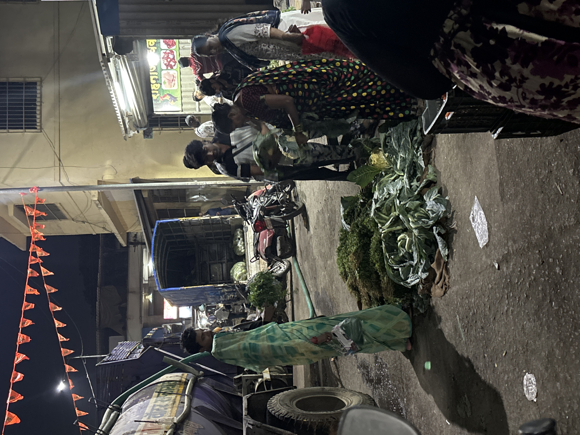 The image depicts a nighttime street scene in Ulhasnagar, India, characterized by active commerce and artificial illumination. The street surface, appearing as dark, damp asphalt, is central to the activity.
In the midground, several large piles of green vegetables, including leafy greens and cauliflower heads, are spread directly on the ground. A woman in a black sari with multi-colored polka dots bends over these vegetables, actively engaging with them. Nearby, a young man in a black and white striped t-shirt observes the scene. To the left, another individual in a light green patterned garment stands beside a large dark blue tanker truck, which has yellow Hindi script (partially visible, likely related to water services). A green hose is visible extending from near the truck to the ground.
On the right side of the frame, a cream-colored building rises, featuring a barred window on an upper floor. Below, a brightly lit shop with a corrugated metal awning displays a sign with Hindi text, prominently featuring "राहनाज़ नॉन-वेज" (Rahanaaz Non-Veg) alongside images of chicken and meat products. Multiple people are visible inside and in front of this shop, including a man in a red and white striped shirt and a woman in a white garment holding a red bag.
Vehicles present include the aforementioned tanker truck, a red motorcycle parked in the midground, and a dark blue mini-truck or tempo loaded with produce. Overhead, against the dark night sky, several strands of orange triangular pennants are strung across the street. Scattered litter, including plastic bags, is visible on the ground. The immediate lower right foreground shows the back of a person next to stacked black plastic crates. The scene indicates a busy market environment operating after dark.