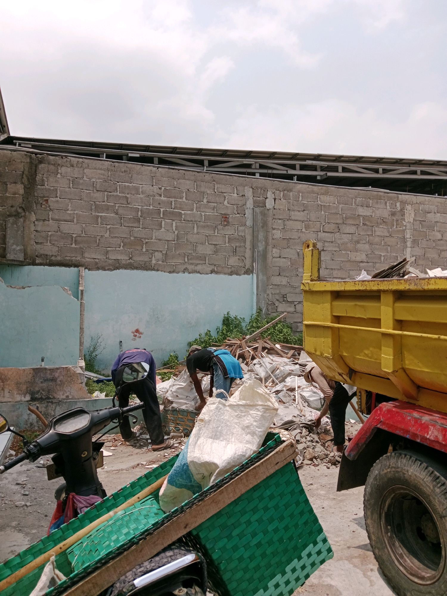 In Cemani, Indonesia, a construction site is depicted with debris scattered across the ground. A yellow dump truck is parked with its bed filled with construction materials. Three men are actively engaged in clearing the debris. One man, wearing a blue shirt and black pants, is bending down and appears to be picking up materials. Another man, in a dark purple shirt and black pants, is also bent over, facing away from the camera. The third man, dressed in a light brown shirt and dark pants, is partially visible behind the dump truck, also engaged in the work. In the foreground, a black motorcycle is parked, and a green woven basket is placed on its side, containing various items. The background features a gray brick wall, indicating the presence of a building. The sky is overcast, suggesting cloudy weather.
