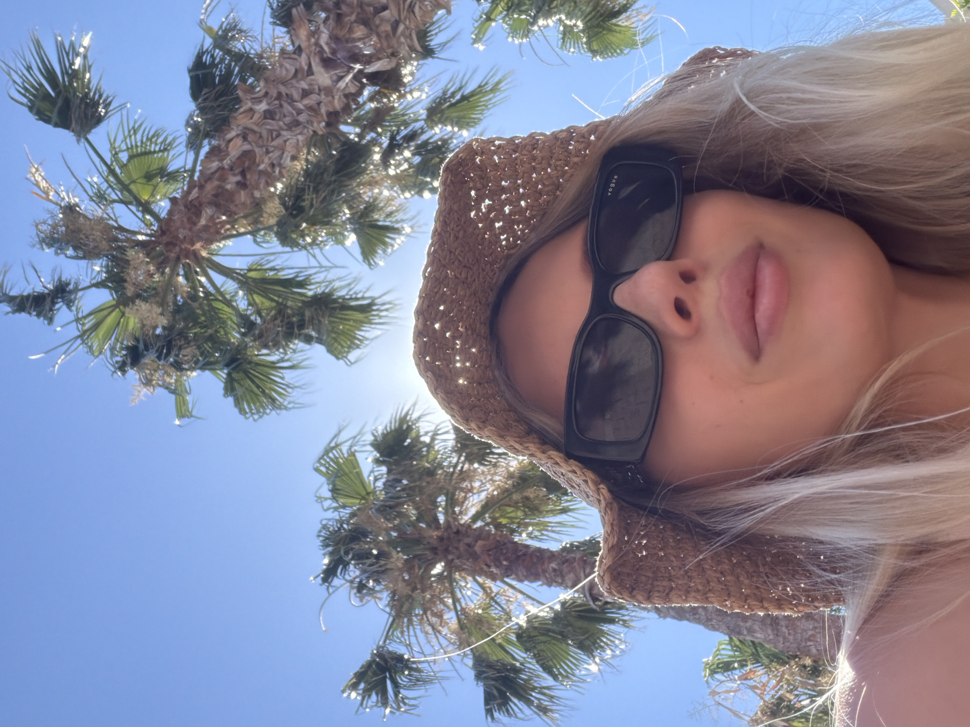 The image depicts a close-up, low-angle shot of a woman wearing a straw hat and sunglasses, positioned in the foreground. Her blonde hair is visible, framing her face. Behind her, several palm trees are silhouetted against a bright blue sky, suggesting an outdoor, warm-weather location. The sun is shining brightly, creating lens flare and highlights on the palm fronds and the woman's hair. The scene appears to be set in Budva, Montenegro, a coastal city known for its beaches and palm-lined promenades.