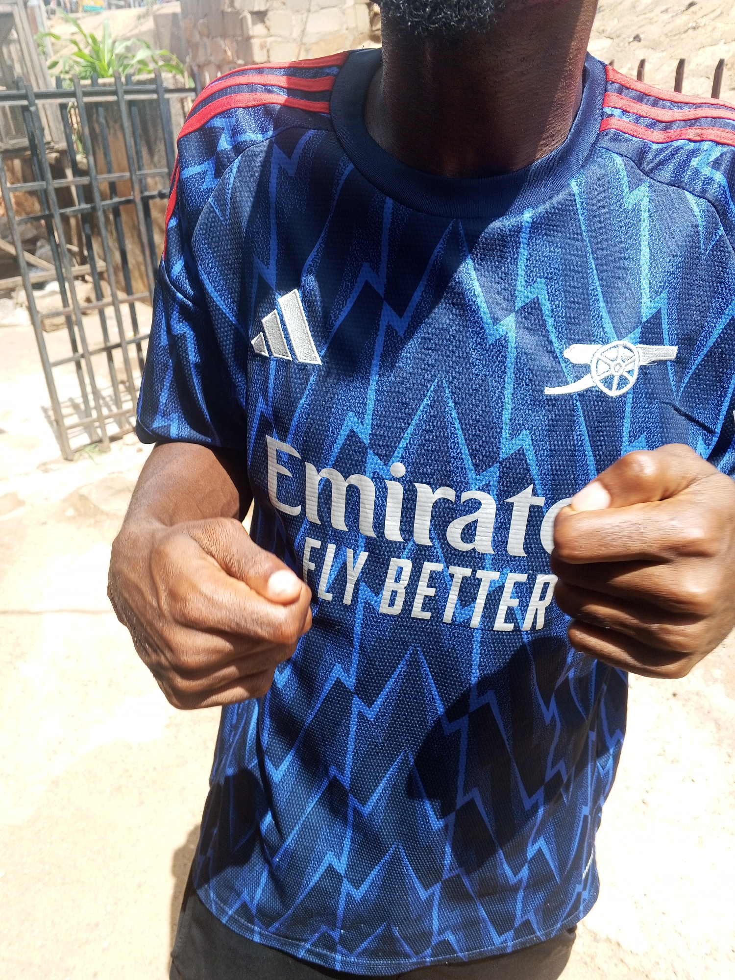 A man is wearing a blue Arsenal soccer jersey with white logos and red shoulder stripes outdoors. The jersey displays "Emirates FLY BETTER" in white lettering. The man has dark skin and a beard. He is standing with his fists clenched. The environment appears to be a residential area in Gwong, Nigeria, as requested. The backdrop includes a fence and possibly some construction.