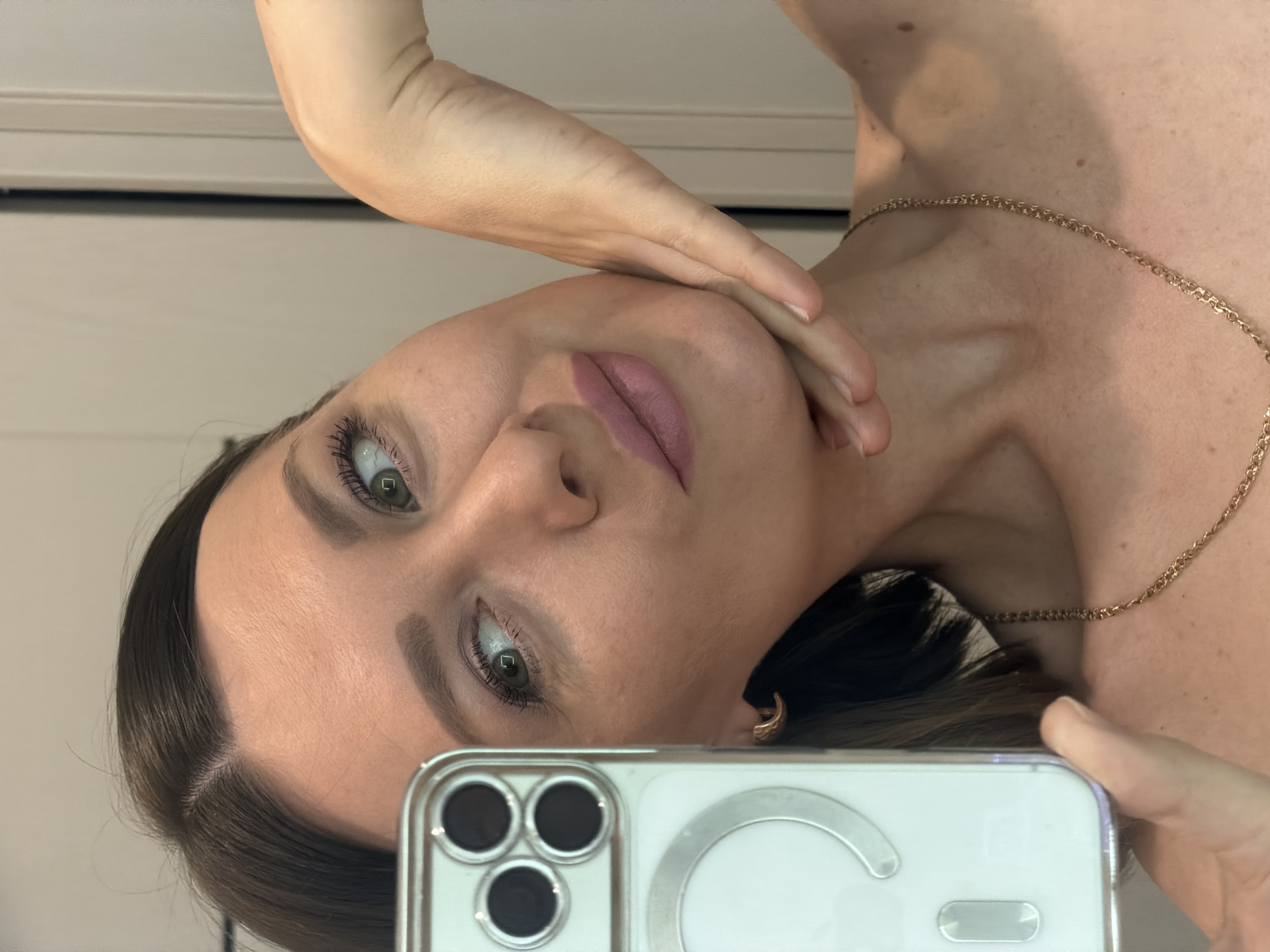 The image shows a selfie of a woman with fair skin, dark hair, and green eyes. She is wearing a light pink lip color and a gold necklace. Her face is partially obscured by her hand and the smartphone she is using to take the picture. The phone has a clear case. The background is a plain, light-colored wall. There is no indication of the location being Rostov-on-Don, Russia.