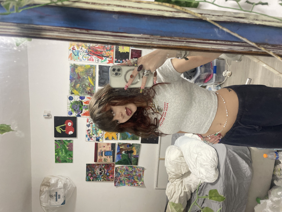 An indoor scene depicts a young female individual taking a mirror selfie within what appears to be a private residence in Raanana, Israel. The female has dark brown hair with reddish highlights and bangs. She is wearing a light grey crop top with the reversed text "TRUST NO MAN. FEAR NO BITCH!" visible in the reflection, and dark blue sweatpants. A pearl and bead chain is draped around her waist. She wears numerous rings on both hands, including a prominent reddish-brown stone ring, and has long, multi-colored fingernails. Her left hand, visible in the reflection as her right, holds a silver-colored smartphone, pointed towards the mirror. A small tattoo is visible on her lower left abdomen. Her tongue is slightly extended.
The background features a white wall extensively covered with numerous small, varied artworks, including abstract designs, a black canvas with two red cherries, a skull motif, a house with a blue door, and a sunflower. To the left, an unmade bed with grey sheets and white pillows is visible. Above the bed, a white plastic bag or cover is affixed to the wall. A green plant vine hangs decoratively in the upper left and right portions of the frame. On the floor, a plastic water bottle with an orange cap is present. A clear plastic storage bin is partially visible in the lower right background. The mirror itself has a distressed, possibly blue or dark-colored, wooden frame.