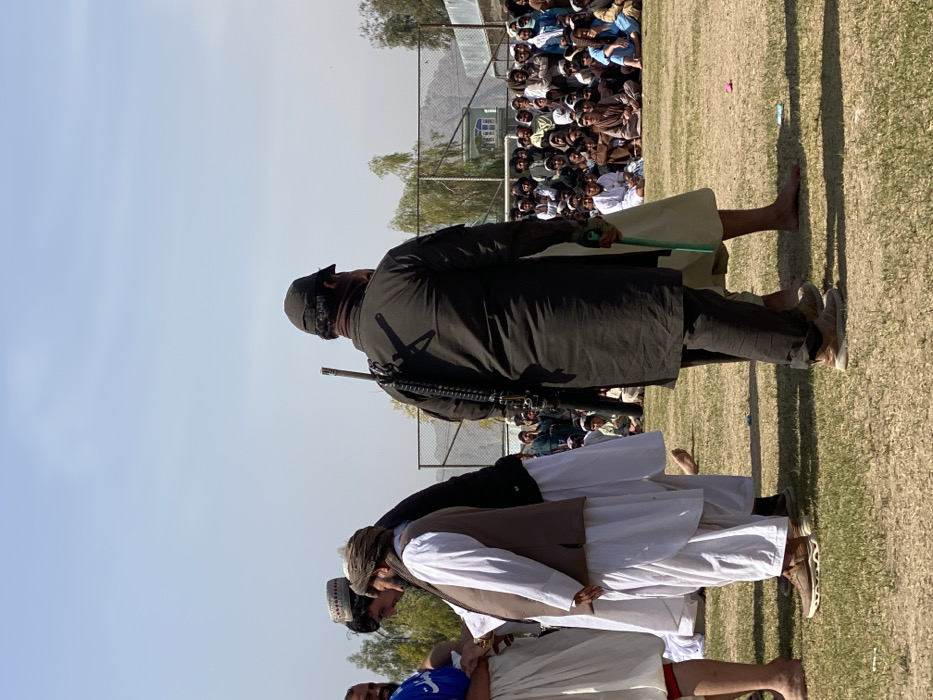A group of men stand on a grassy field, likely spectators. One man stands out, wearing dark clothing with a large rifle slung over his shoulder. A second man in traditional garb is barefoot. A chain-link fence is visible in the background, and behind it a larger crowd of people can be seen observing from a distance. The sky is clear and the sun is out.