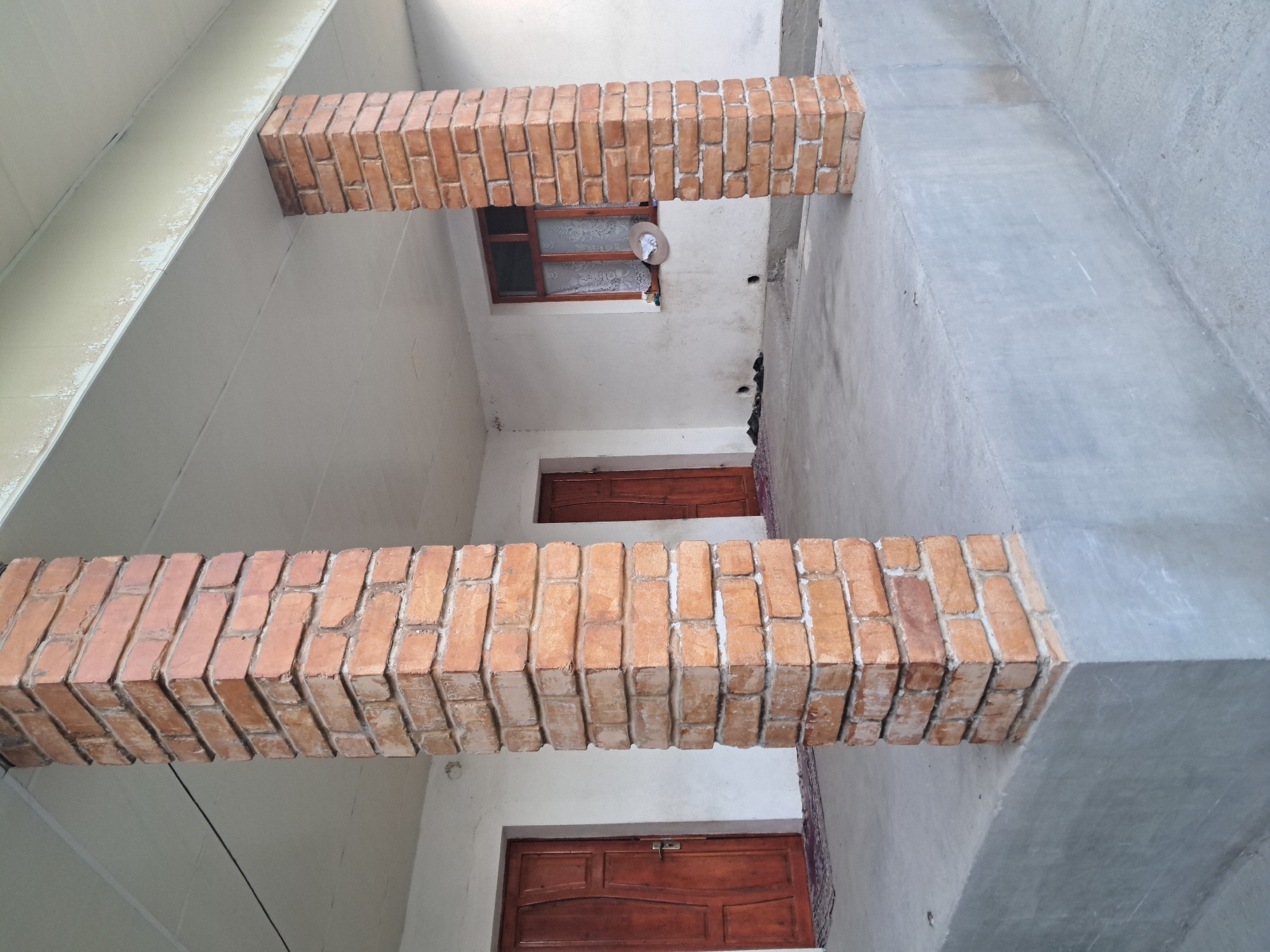 The image shows a porch area of a building. Two brick pillars, constructed from reddish-brown bricks, support a white ceiling. To the left, there is a wooden door with a metal handle. To the right, a window with wooden frames and white lace curtains is visible. In front of the pillars, a concrete platform is raised. The ground outside the platform appears to be paved. The environment suggests a residential setting in the city of Bagish, Uzbekistan.