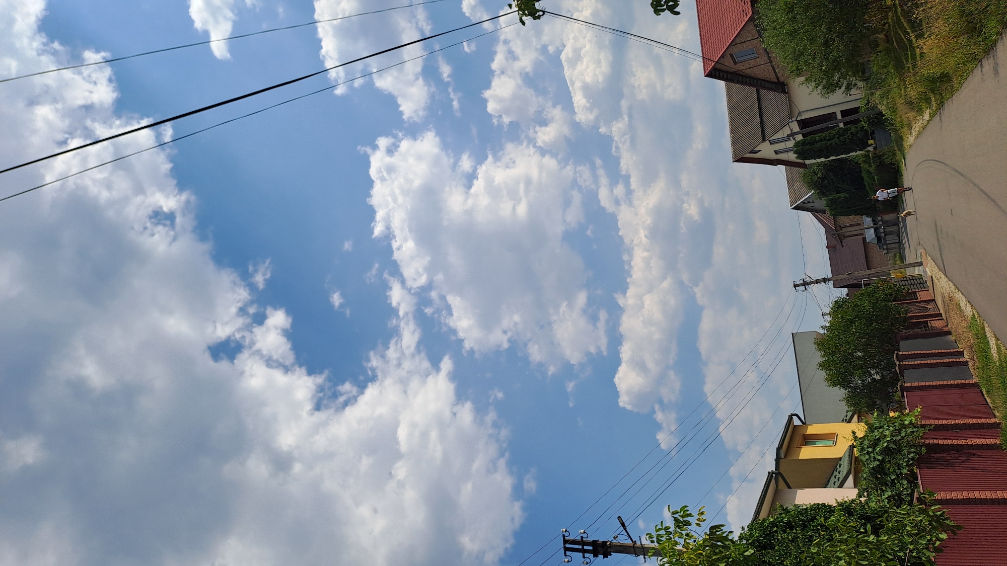 The image depicts a suburban street scene in Kolomyia, Ukraine, under a partly cloudy blue sky. A paved road runs through the center of the frame, lined with residential houses and fences. On the left, a multi-story house with a yellow facade and green roof is visible, bordered by a tall red metal fence and lush green foliage. Further down the street on the left, there is a utility pole with wires. On the right side of the road, several houses with pitched roofs are visible, interspersed with trees. In the distance, a person is seen riding a scooter, accompanied by a dog on the road. Overhead, power lines crisscross the sky. The overall impression is a quiet, sunny day in a residential area.