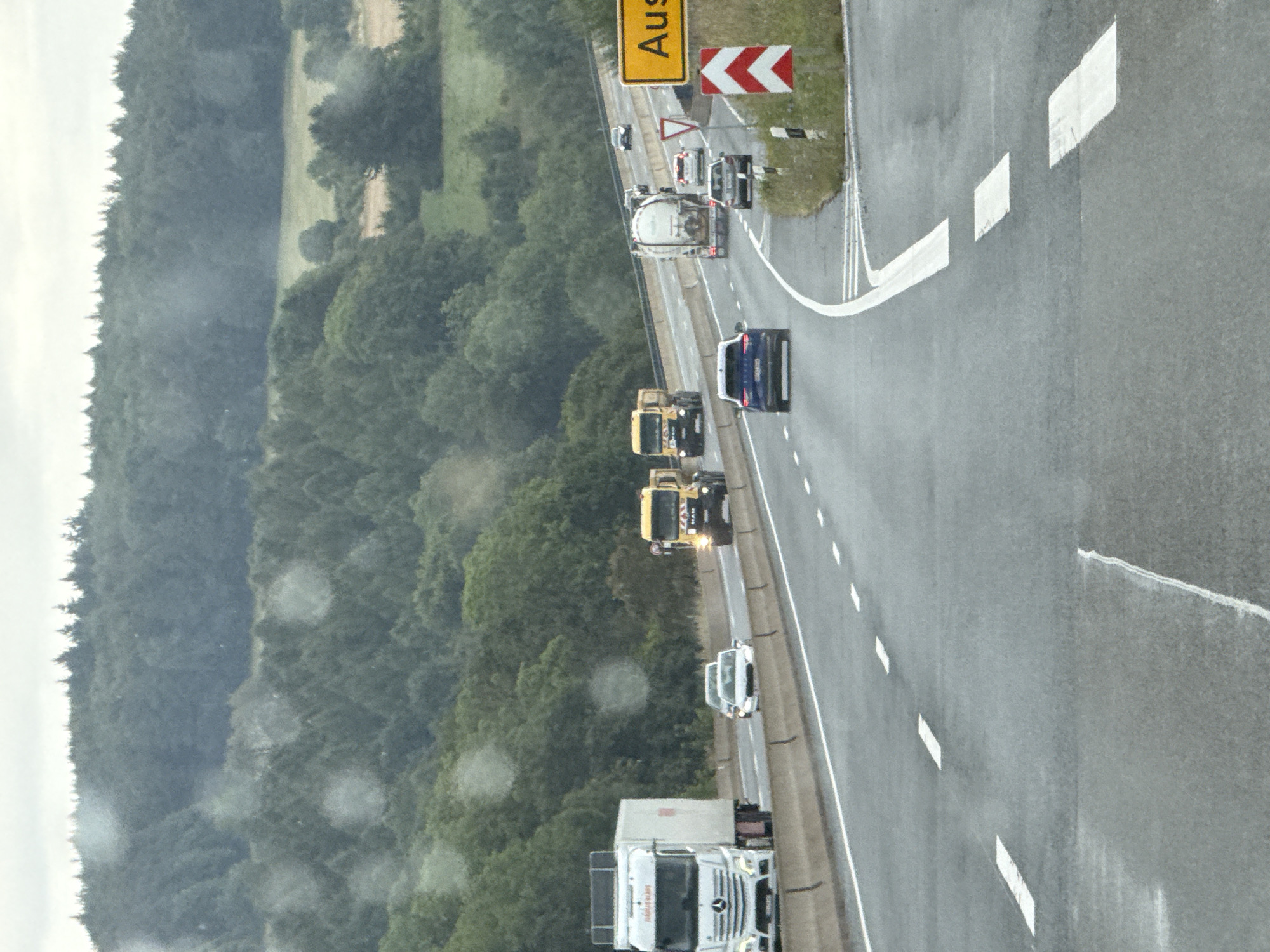 The image captures a view of a multi-lane highway in or near Bad Lauterberg im Harz, Germany, under an overcast sky. A white Mercedes-Benz semi-truck with a white trailer is traveling in the left lane, while a white passenger car is in the right lane behind it. Further down the highway, a yellow MAN truck with flashing lights is in the right lane, followed by a white tanker truck and a white passenger car. A blue SUV is in the left lane. The highway is bordered by a concrete barrier on the left and a grassy embankment with trees on the right. A yellow road sign with the word "Aus" is visible on the right side, along with a red and white chevron marker indicating a curve or hazard. The road surface appears wet, suggesting recent rain.