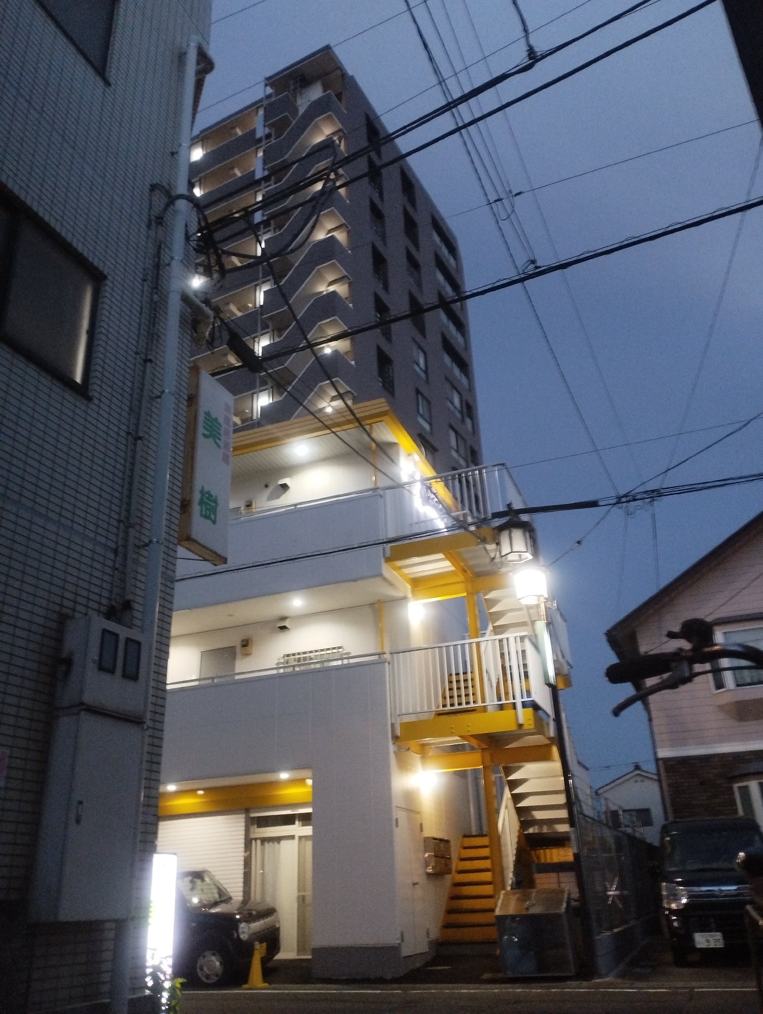 In the city of Fussa, Japan, on a cloudy evening, an apartment building stands tall. The building has multiple stories with gray exteriors and balconies. Yellow accents are visible on the balconies and an outdoor staircase. A sign with Japanese characters is hanging on the side of a building in the foreground. Two cars are parked near the apartment building. The sky is a deep blue, indicating dusk. Power lines are strung across the sky.