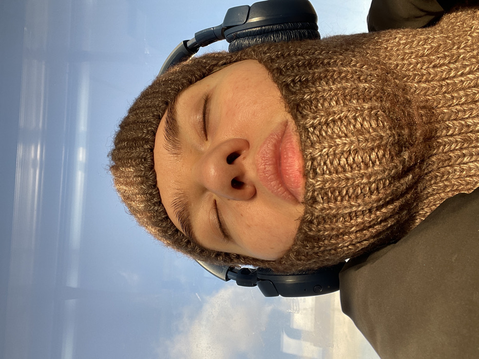 A close-up, low-angle photograph shows an individual from the upper chest upwards, with eyes closed. The subject wears a light brown, ribbed knit balaclava covering the head, neck, and lower face, exposing only the nose and mouth area. Dark-colored over-ear headphones are worn over the balaclava. The individual's skin appears warm-toned, illuminated by natural light. A portion of a dark-colored outerwear garment is visible on the lower left. The background features a clear blue sky with sparse white clouds, visible through a window that exhibits horizontal reflections, indicating an interior perspective from a vehicle. This scene is located in Moscow, Russia.