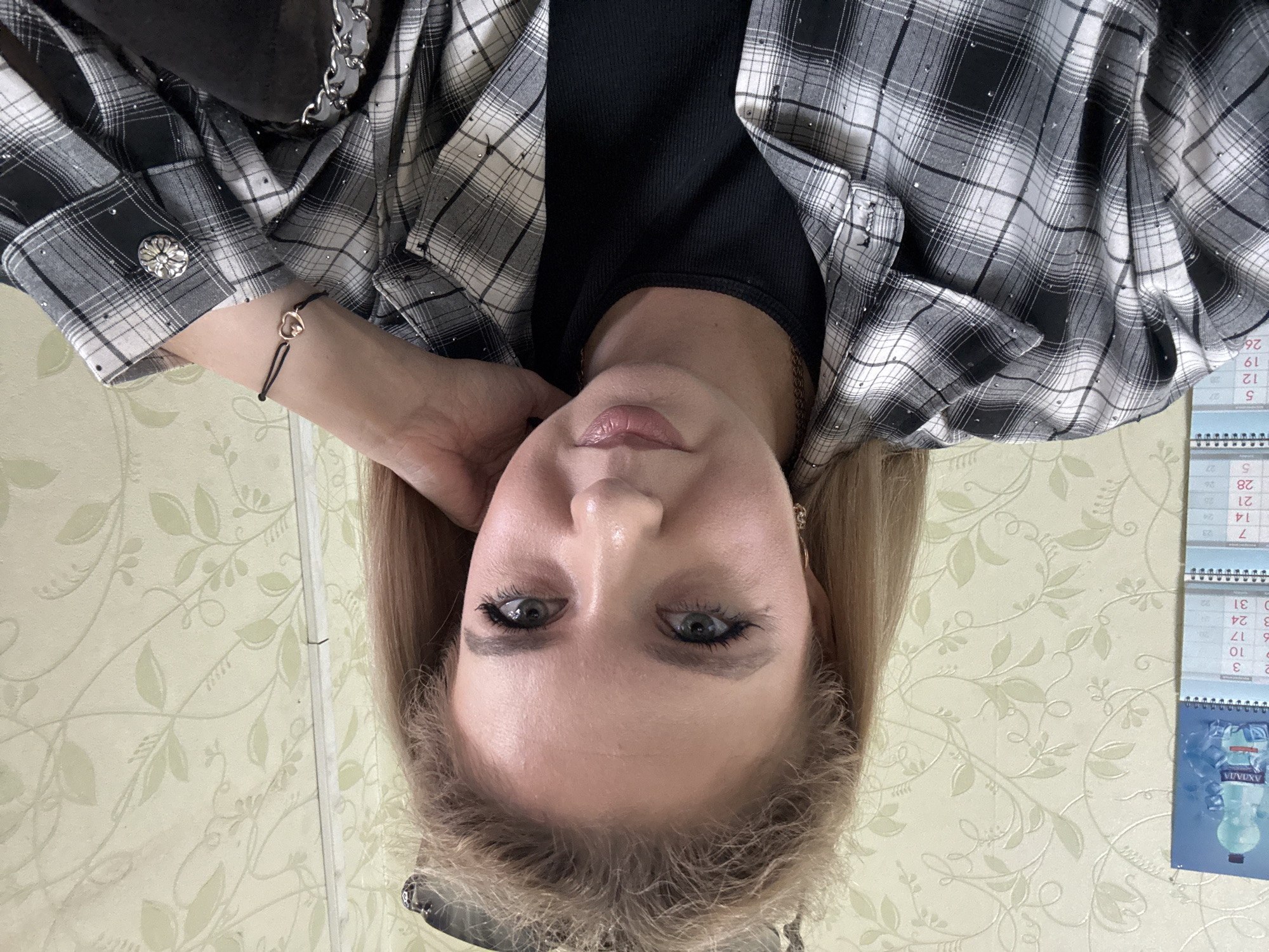 The image shows a close-up, selfie-style shot of a young Caucasian woman. She has long, blonde hair, light-colored eyes, and appears to be wearing makeup, including eyeliner and mascara. She is wearing a black ribbed top under a grey and white plaid shirt adorned with small, reflective embellishments. A delicate bracelet with a heart charm is visible on her left wrist. She is resting her right hand on her neck and chin, and her left arm is bent. In the background, a calendar with numbers and some text, as well as a bottle of water, are visible on the left side. The wall behind her has a light-colored floral wallpaper pattern. The lighting is moderate, suggesting an indoor setting. No specific city or location details are discernible from the image.