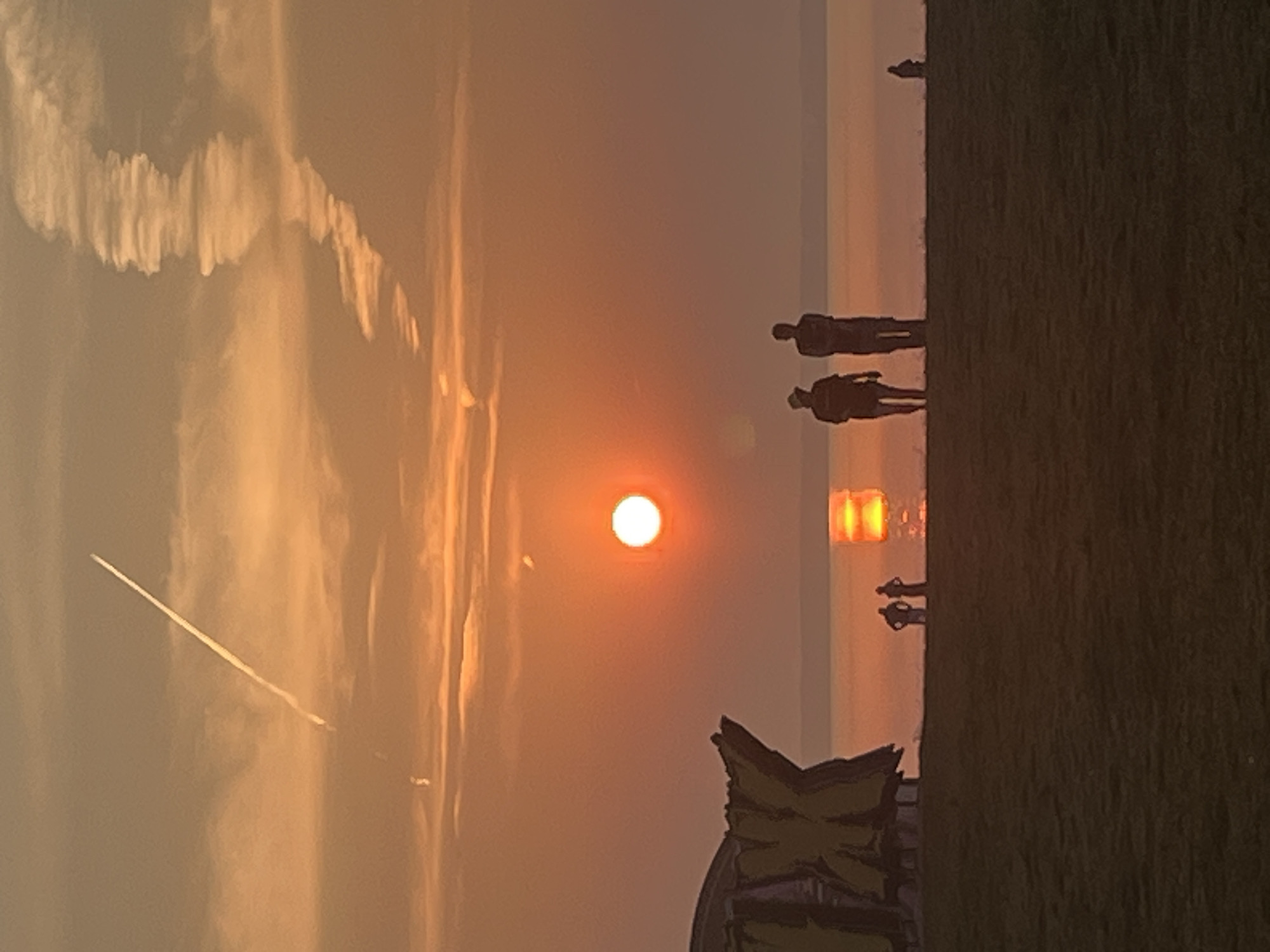 The image depicts a sunset over a body of water, possibly the sea, with a faint orange glow illuminating the sky. In the foreground, there is a grassy field. To the left, a partially visible, abstract, yellow sculpture is present. In the middle ground, silhouetted against the setting sun, are several figures standing on a shoreline. Two of these figures are taller and appear to be statues or monuments, while two smaller figures are standing near them. Further to the right, a single, very small, silhouetted figure is visible near the water's edge. The sun is a prominent, bright orange disc low on the horizon, reflecting a warm glow on the water. Two distinct contrails are visible in the sky, one long and thin, the other a more diffused, wispy formation. The overall scene conveys a sense of tranquility and natural beauty. The location is specified as Surakovo, Russia.
