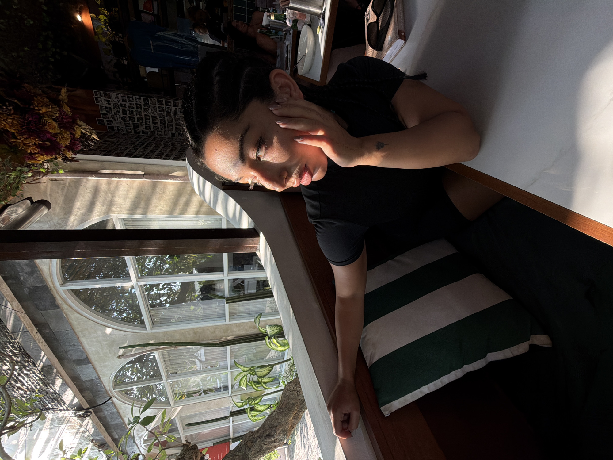 A young woman with dark braided hair is seated at a table in an outdoor cafe in Mengwi, Indonesia. She is wearing a black t-shirt and resting her right hand on her cheek, looking to her left. Sunlight streams in from the left, illuminating her face and casting shadows. Behind her, through a large arched window, a lush green garden with plants and trees is visible. The cafe interior includes a wall adorned with many small framed photographs and hanging dried flowers. On the table in front of her are plates, glasses, and a book. The bench she is sitting on is covered with a cushion featuring green and white stripes.