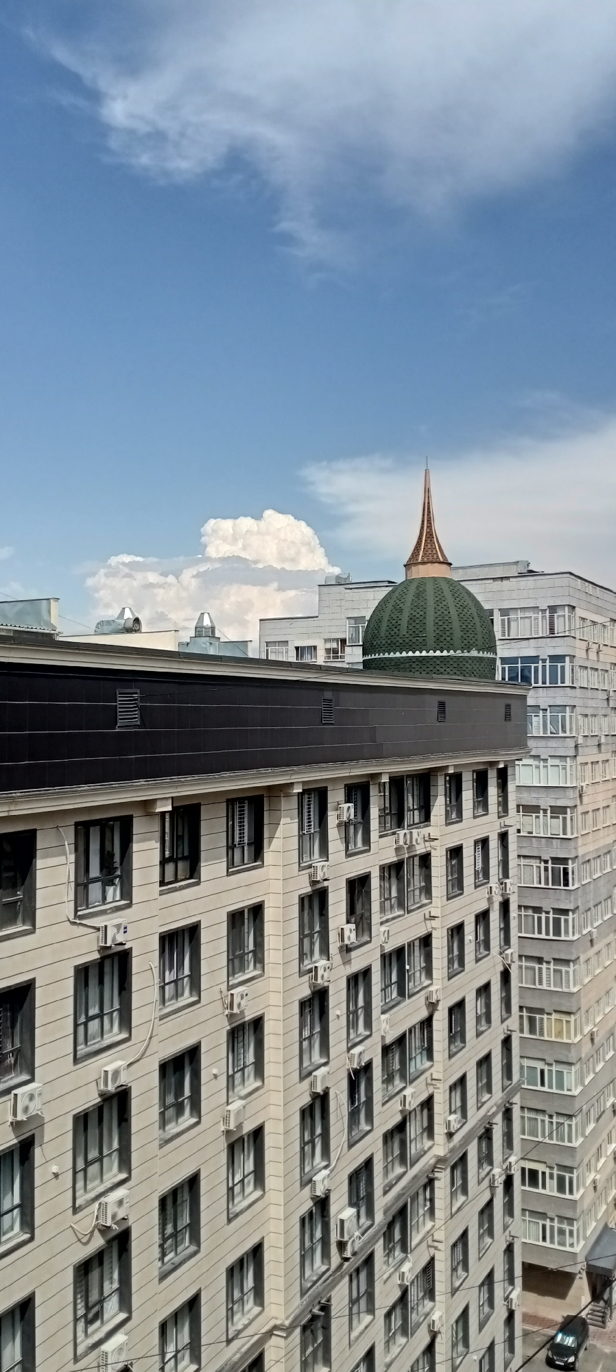 The image shows a view of apartment buildings in Bishkek, Kyrgyzstan, under a partly cloudy sky. The foreground features a light-colored building with numerous windows and air conditioning units mounted on its facade. Above this building, a dark, flat roof is visible. In the background, a taller, gray apartment building with many windows can be seen to the right, and further to the right, a green domed structure with a golden spire rises above other buildings. A large, puffy white cloud dominates the upper left portion of the sky, with smaller wisps of clouds scattered throughout the blue sky. The scene is static and captures an urban environment.