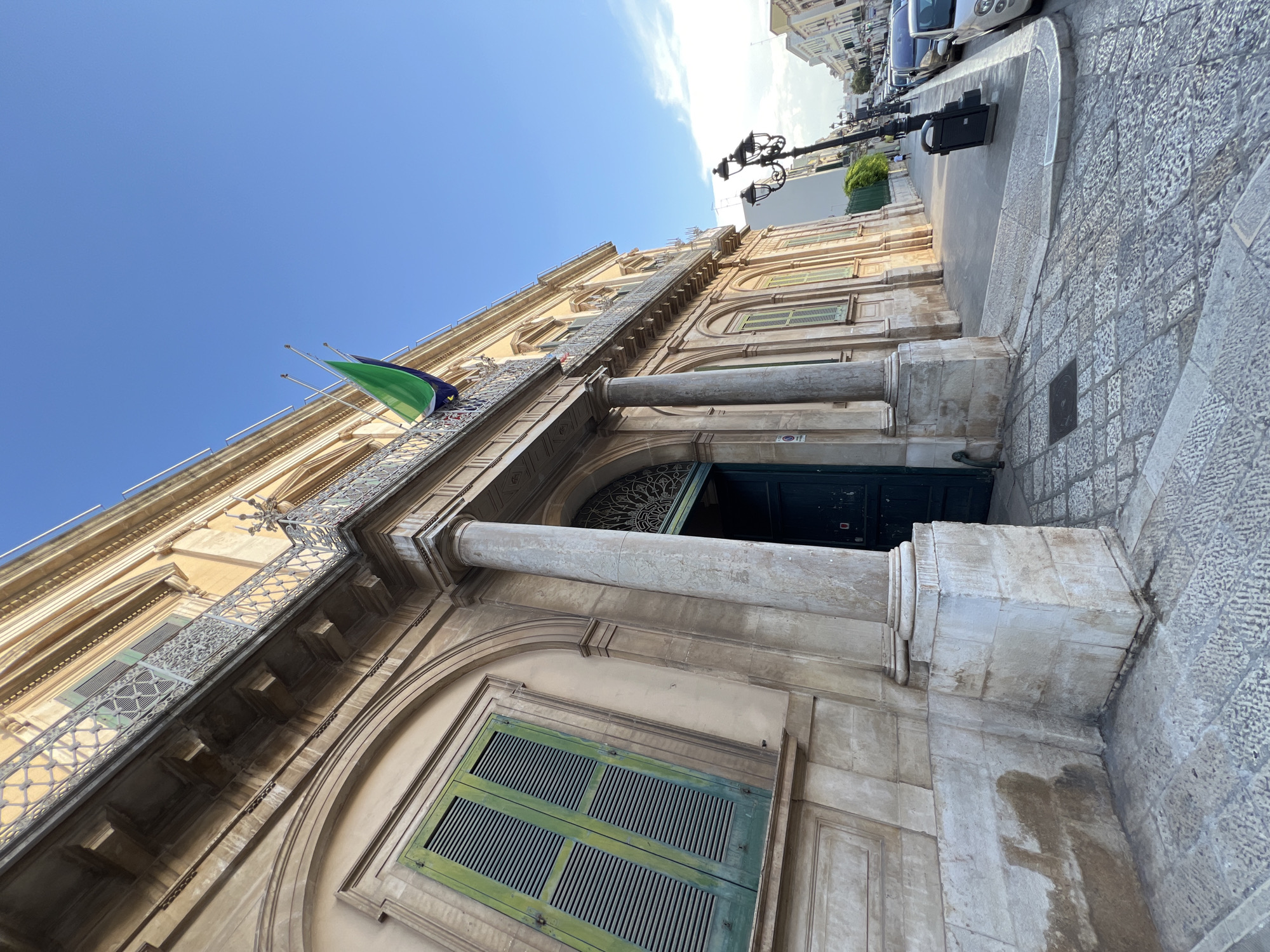 The image captures a low-angle view of the facade of a historic building, likely the Palazzo Comunale in Bitonto, Italy. The building exhibits classical architectural features, including large stone columns, arched windows with green shutters, and decorative balconies with intricate metal railings. A flag, predominantly green with a dark blue section, is visible flying from a balcony. The street in front of the building is paved with cobblestones, and a traditional street lamp stands to the right. Cars are parked along the street, and a few people are visible in the distance, walking on the sidewalk. The sky is clear blue with a few wispy clouds. The overall scene suggests a sunny day in an Italian city, highlighting the architectural heritage of Bitonto.