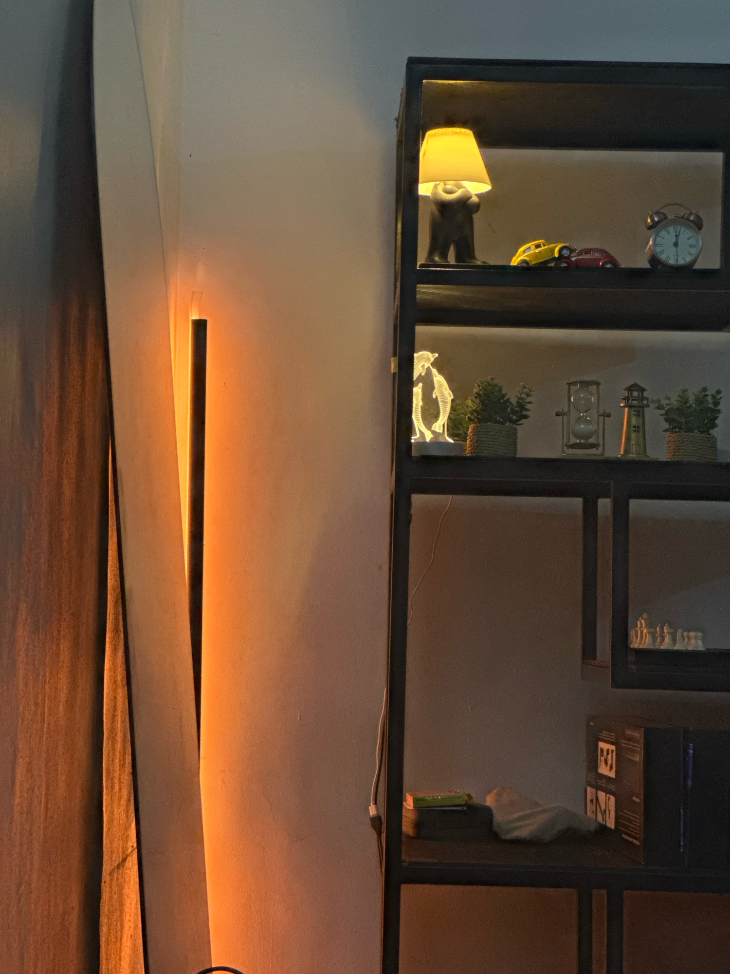 The image depicts an indoor scene featuring a tall, black, metal shelving unit against a wall. The shelving unit is illuminated by a vertical, orange-hued light source positioned to the left of the unit, casting a warm glow on the wall and the edge of the shelving unit.
The top shelf of the unit displays a small, brown lamp shaped like a bear holding up a yellow lampshade, two miniature toy cars (one yellow, one red), and a vintage-style alarm clock. The second shelf from the top features a luminous acrylic sculpture of two dolphins, a small potted plant with green foliage, an hourglass, a miniature lighthouse, and another small potted plant. The third shelf from the top is mostly empty and dark, but a section on the right appears to contain a white chess set. The bottom shelf holds a stack of books, a folded light-colored cloth, and a dark box with some text and graphic elements on its side. A thin white wire runs down the front of the shelving unit.
The wall behind the shelving unit is a pale, neutral color, and a draped curtain or fabric hangs on the far left, partially obscuring the light source. The overall ambiance is dimly lit and cozy, with the warm light from the vertical lamp dominating the left side of the frame. The scene is devoid of people, indicating it is likely a still-life arrangement of decorative objects in a domestic or retail setting. The image provides no specific clues to pinpoint its location within Surulere, Nigeria, beyond being an interior space.