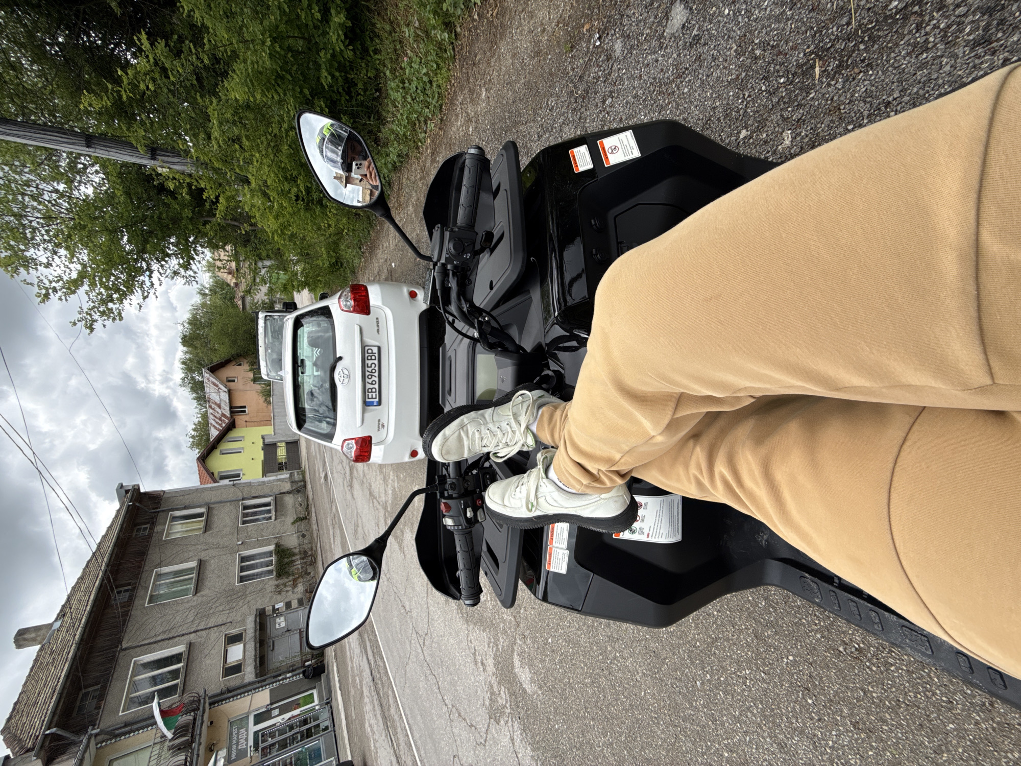 The image depicts a person seated on a black quad bike on a street in the city of Брусовeте, Bulgaria. The person is wearing light-colored pants and white sneakers, with their feet resting on the bike's front section. The quad bike is parked on a paved road next to a white Toyota Auris, which has a Bulgarian license plate number EB 6965 BP. The background shows buildings characteristic of Брусовeте, including a multi-story gray building with windows and a commercial establishment on the ground floor with the sign "МИНИ МАРКЕТ ДИДИ". Other buildings in the background include a yellow and white house with a red roof. The sky is overcast. The quad bike's mirror reflects the person holding a phone.