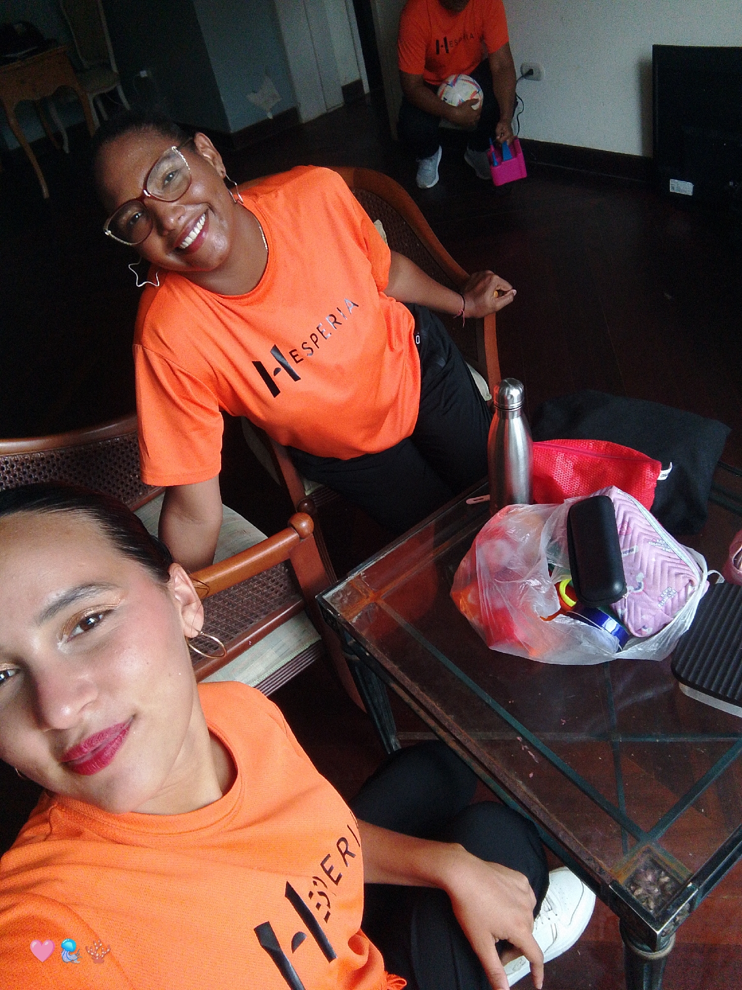 Two women are in a room, likely a living room or common area, given the presence of a couch, wooden furniture, and a dark wooden floor. The lighting suggests it is daytime, possibly late morning or afternoon.
In the foreground, a woman wearing an orange t-shirt with the word "HESPERIA" visible on it is looking directly at the camera and smiling. She has dark hair pulled back and is wearing glasses with a decorative, angular frame. Her t-shirt has a graphic of a stylized "H" and "ESPERIA" written in black.
In the background, another woman is also wearing an orange "HESPERIA" t-shirt and is crouching, holding a soccer ball and a pink pump. She appears to be preparing for some activity.
On a glass-top coffee table between the two women, several items are laid out: a silver water bottle, a red mesh bag, a clear plastic bag containing various colorful objects including what looks like a pink makeup bag or pouch, and a black electronic device.
The overall mood seems relaxed and casual, possibly indicating a break or preparation for a recreational activity. The presence of the soccer ball and pump suggests a sports-related event or practice. The setting appears to be indoors.
