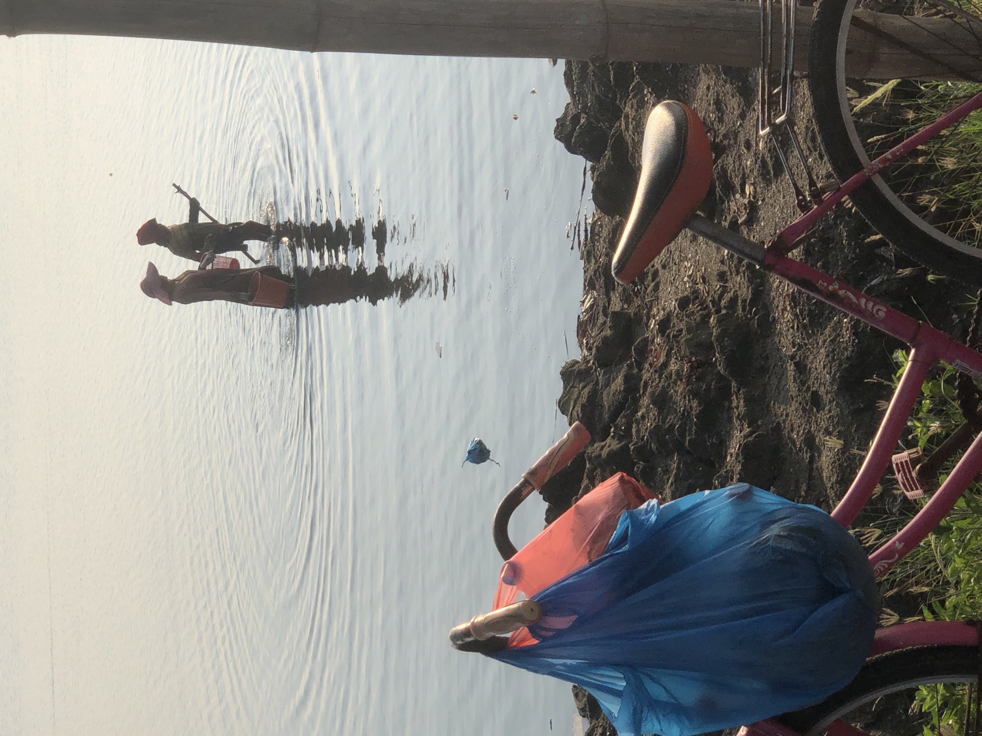 Two individuals, appearing to be women, stand in shallow water near the shore of the Krueng Cunda River in Lhokseumawe, Indonesia. Both are wearing wide-brimmed hats. The person on the left, facing right, holds a red bucket. The person on the right, facing left, holds a stick and appears to be fishing or collecting something from the water. They are both submerged up to their knees. In the foreground, a pink bicycle is parked on the muddy bank. Blue and red plastic bags are tied to the handlebars, and the bicycle is laden with items. The water is calm, with subtle ripples spreading outwards from the individuals. The bank is composed of dark, clumpy soil and sparse vegetation. A vertical wooden pole stands to the right of the frame, casting a reflection on the water's surface. The scene is bathed in a soft, warm light, suggesting it is either early morning or late afternoon.