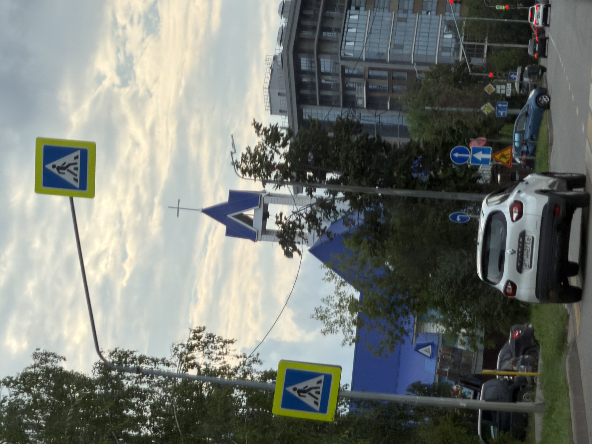 The image captures a street scene in Kazan, Russia, characterized by a cloudy sky and a mix of architectural styles. In the foreground, a white car drives away from the camera, with other vehicles, including a dark SUV and a blue hatchback, visible on the road and parked. Traffic signs, including pedestrian crossing symbols and directional arrows, are present along the roadside. Beyond the road, a modern multi-story building with glass facades stands adjacent to a structure with a blue roof and a prominent cross on its steeple, suggesting a church or religious institution. Trees and greenery are interspersed throughout the scene, contributing to the urban landscape. The overall impression is of a typical day in a Russian city, with active traffic and varied architectural elements under a diffused sky.