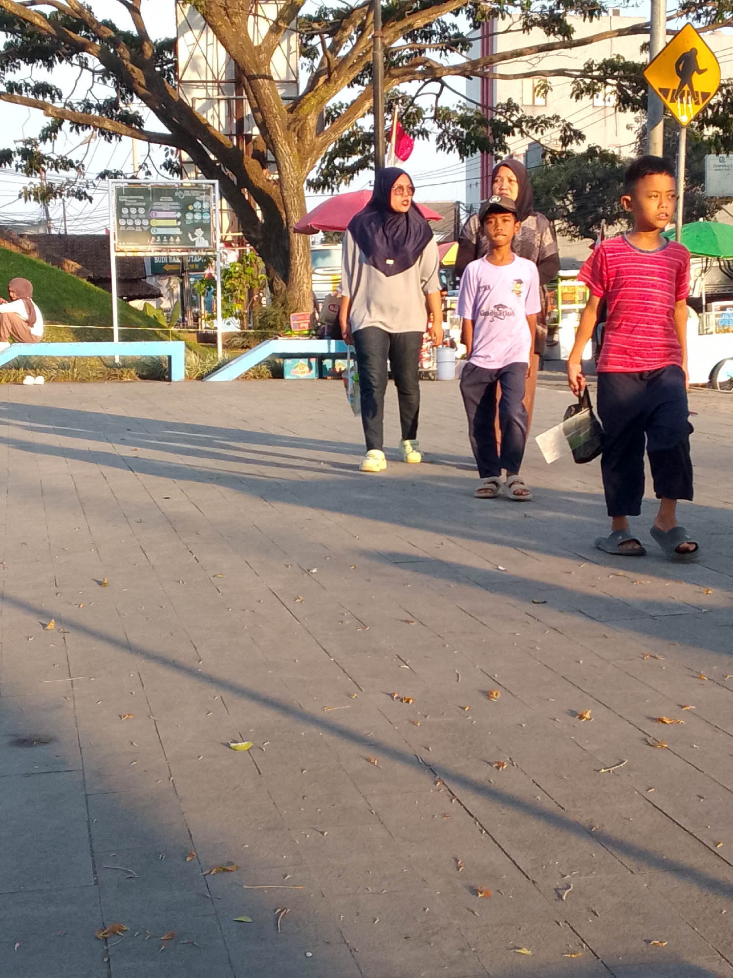 In Ciburuy, Indonesia, four individuals are walking on a paved walkway. The group consists of two adults and two children, all appearing to be of Indonesian descent. On the left, a woman wearing a dark blue hijab and a gray shirt walks with a teenage boy in a light purple shirt and dark pants, and sandals. To their right, another woman, also wearing a dark hijab, walks behind them, partially obscured. A boy in a red and pink striped shirt and dark pants walks on the far right, carrying a dark bag. The walkway is shaded by a large tree with a thick trunk and branches, casting long shadows across the gray paved surface. In the background, there is a grassy slope, some trees, and buildings. A yellow pedestrian crossing sign is visible on a pole to the right of the group. The scene appears to be an outdoor public space, possibly a park or a promenade, bathed in the warm light of what seems to be either sunrise or sunset.
