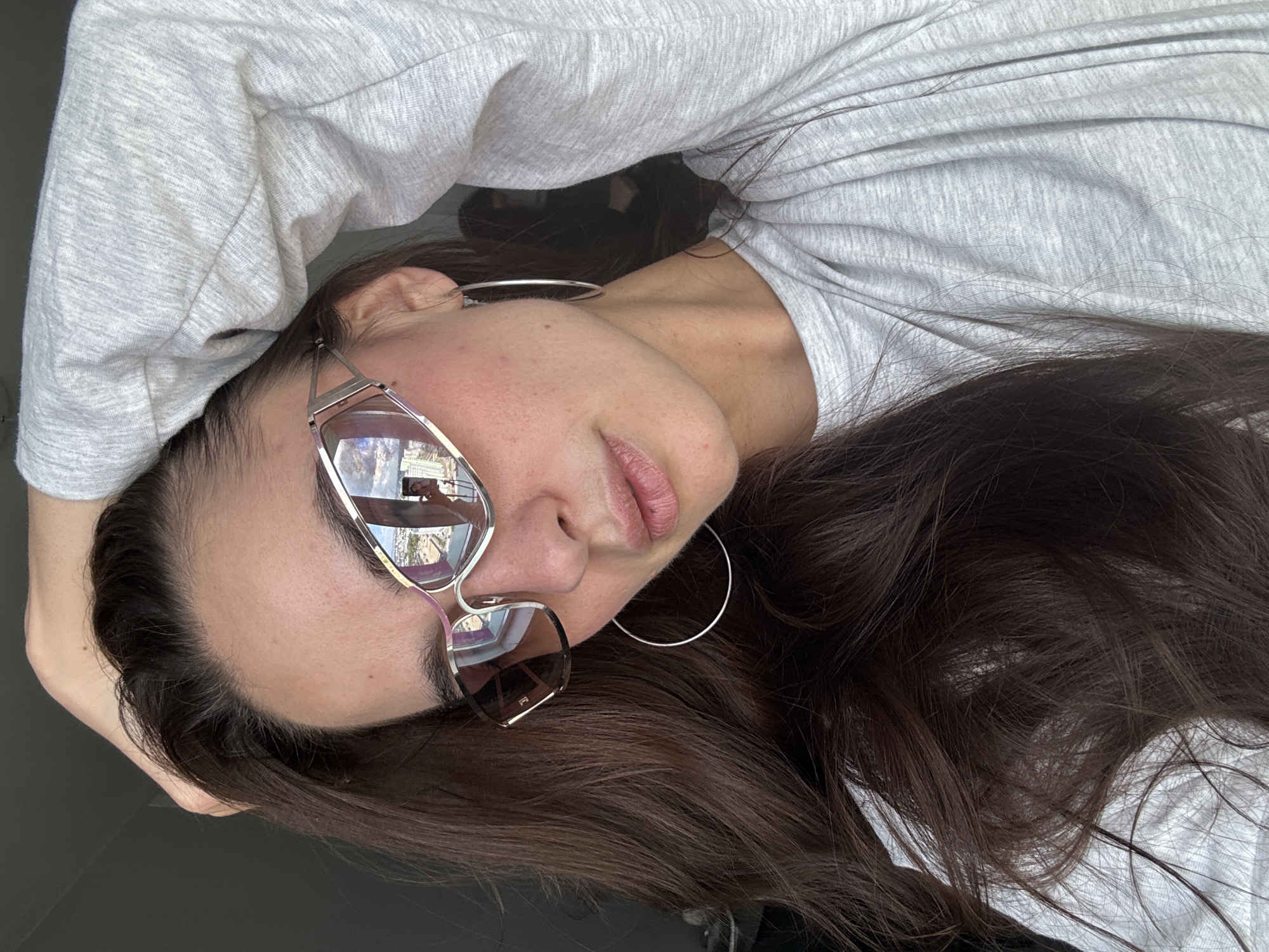 A woman with brown hair is lying on her back wearing a light grey long-sleeved shirt and silver hoop earrings. She is also wearing silver framed sunglasses that reflect a cityscape with a river and buildings, likely the Yenisey River and parts of Krasnoyarsk, Russia. The background is a dark grey surface.