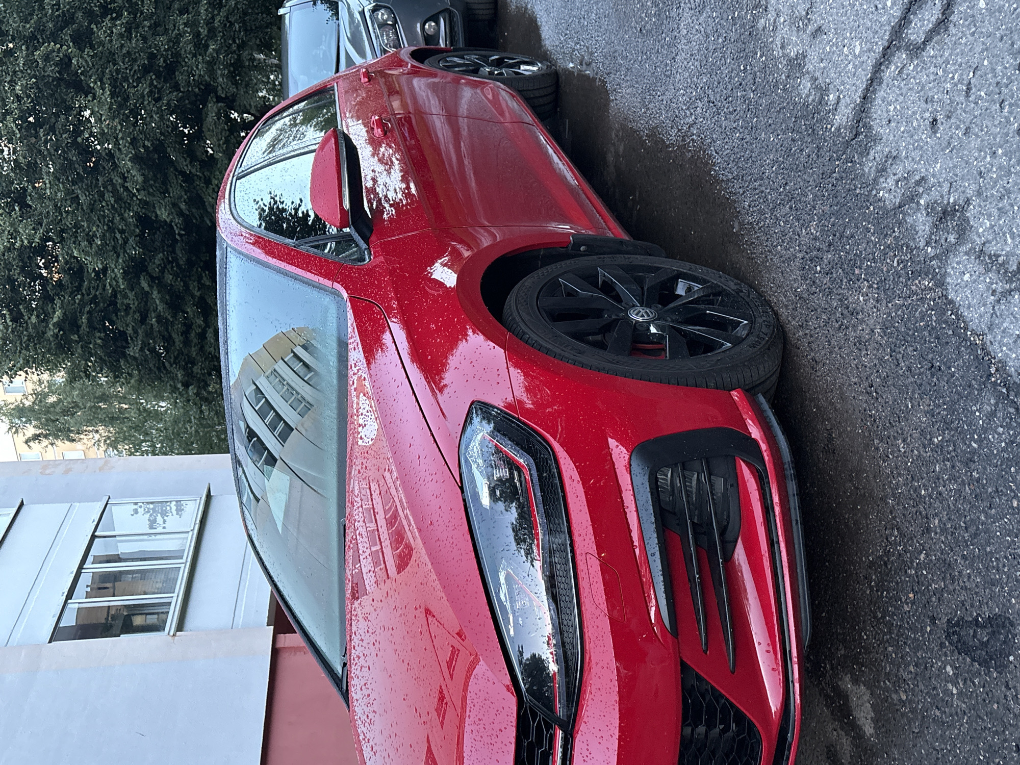 The image shows a close-up, low-angle view of a red Volkswagen Golf GTI parked on an asphalt road. The car is wet, with numerous water droplets visible on its surfaces, suggesting it has recently rained or the car is being washed. The front driver's side wheel, with its black alloy rim and Volkswagen logo, is prominently featured. The car's headlight, with its distinctive LED daytime running lights, and a portion of the front bumper and grille are also visible. The background is blurred, but a building with windows and a lush green tree can be discerned. Another vehicle, likely a dark-colored car, is parked behind the red Golf. The lighting suggests it is daytime, possibly overcast due to the wet conditions and diffused light. The scene appears to be in a residential or urban area, given the presence of the building and the parked cars. There is no visible text in the image.