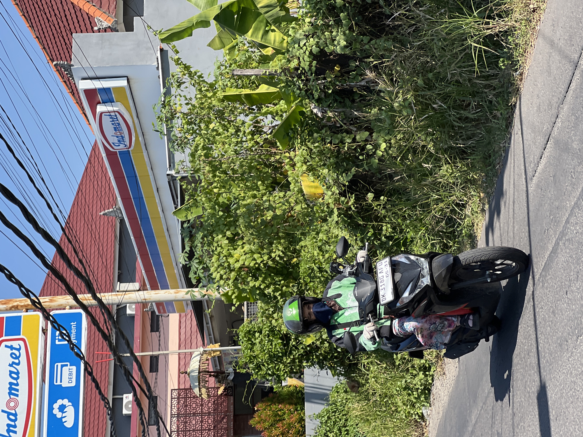 A person wearing a green jacket and helmet, with a blue mask covering their face, rides a black scooter on a paved road. The scooter has a license plate that reads "DK 318G AFU 05-31". The person is carrying a bag on the back of the scooter. The background shows lush green vegetation, a building with red tile roofs, and a sign that says "Indomaret", a convenience store chain common in Indonesia. The sky is clear and blue. The scene appears to be in Denpasar, Indonesia, a city known for its vibrant culture and natural beauty.