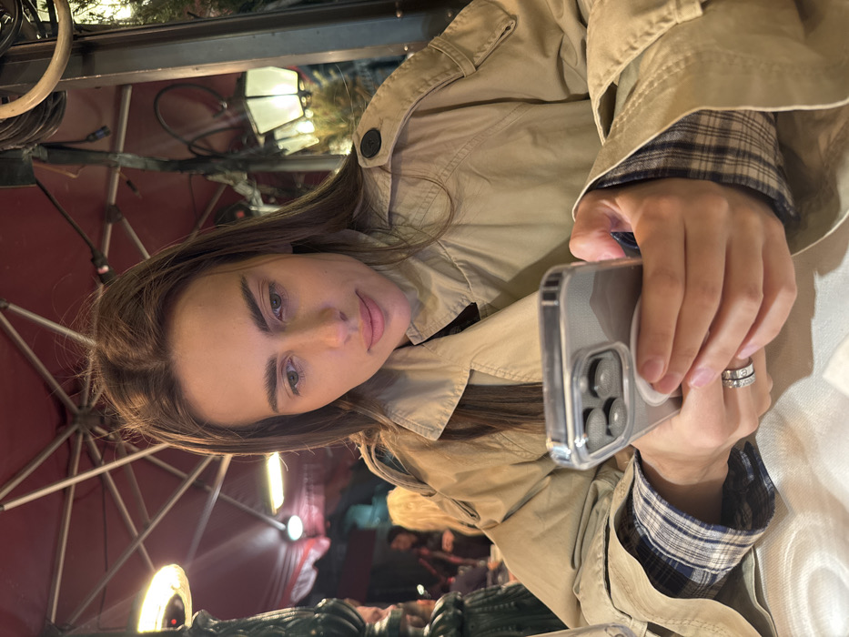 An outdoor evening scene depicts a female seated at a table in what appears to be a restaurant or café in Venice, Italy. The subject, a woman with long, straight brown hair and a fair complexion, is wearing a beige trench coat over a blue and white plaid shirt. She wears a metallic ring on her left ring finger. Her gaze is directed downward, focused on a smartphone held with both hands. The phone is encased in a clear protective cover and features a white pop socket on its back.
The environment includes a white tablecloth on the table. Overhead, a red canopy or large umbrella structure is visible, along with an operational outdoor heater providing reddish-orange light from the left. A decorative dark pillar stands to the left of the frame. In the blurred background, additional patrons are faintly visible, along with classic street lamps or lanterns emitting warm light, and some green foliage, contributing to the ambiance of an outdoor dining establishment at night.