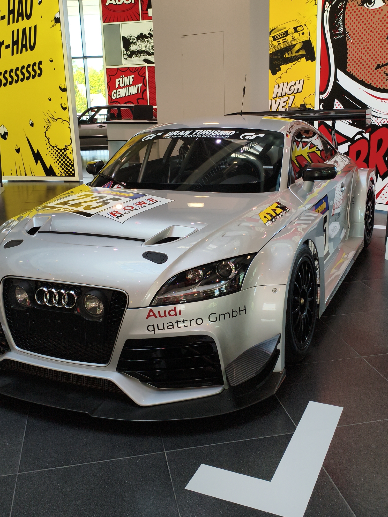 The image depicts a silver Audi TT RS GT4 racing car on display, likely within the Audi Forum in Ingolstadt, Germany. The car is positioned on a dark tiled floor. Behind the car, large, colorful graphic posters are visible. One poster on the left is yellow with black text that reads "HAU-HAU-HAU ssssssss". Another poster to the right is yellow and features a yellow Audi car with the text "HIGH FIVE!". To the far right, a poster with red and white comic-style graphics is visible. The car itself has racing decals, including the number 23 on the hood, and "Audi quattro GmbH" branding on the front bumper. The car's headlights, grille, and large rear spoiler are prominent. The environment appears to be an indoor exhibition space, possibly a museum or showroom, with large windows in the background allowing natural light to enter. There are no people visible in the image.