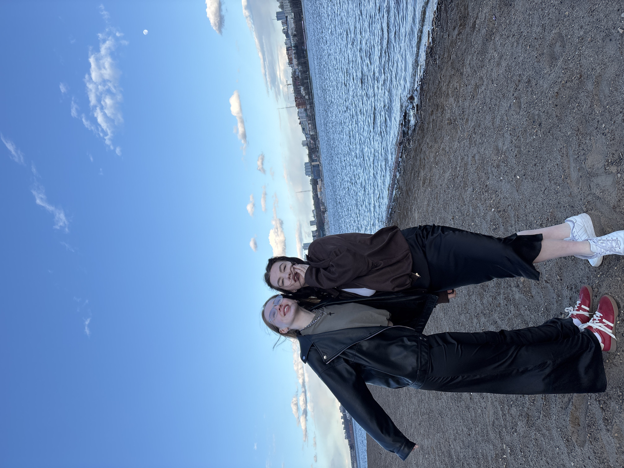 Two young women are captured in a joyful moment, posing for a photograph on a gravelly bank next to a wide body of water, likely a river. The image is oriented vertically, showing a vibrant blue sky with scattered white clouds and a visible moon in the upper left quadrant.
In the foreground, a woman with light brown hair and glasses wears a black leather jacket over an olive-green top, a silver chain with a small cross pendant, and long dark trousers or a skirt, paired with red and white sneakers. She is smiling broadly and looking towards the camera. Slightly behind her and to her right, another woman with dark hair wears a brown jacket over a white top, dark trousers or a skirt, and white sneakers. She is also smiling, with one hand near her face. Both women appear happy and relaxed.
Across the water, an urban skyline is visible, featuring various buildings and what appears to be construction cranes, indicating a city environment. The water itself shows gentle ripples reflecting the daylight. The ground beneath their feet is composed of small stones and sand. The bright conditions suggest it is daytime, possibly late afternoon or early evening given the presence of the moon. There is no visible text in the image.