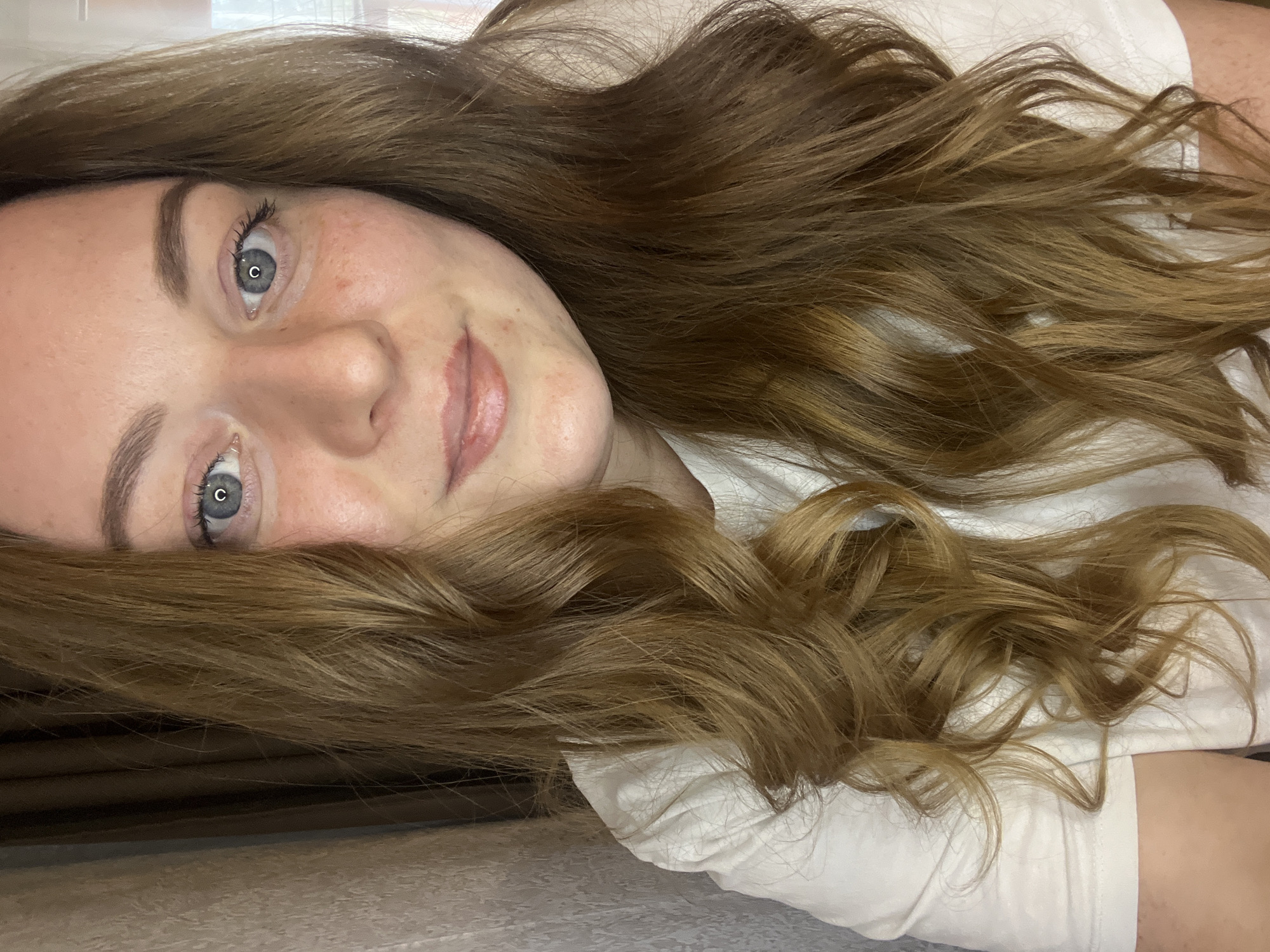 A woman with light skin, freckles, blue eyes, and wavy brown hair is lying down, wearing a white t-shirt. The background consists of a neutral-colored textured surface and a dark brown vertical strip, possibly a wall or curtain. The location is not identifiable as Раздолье, Russia, based on the visual information.