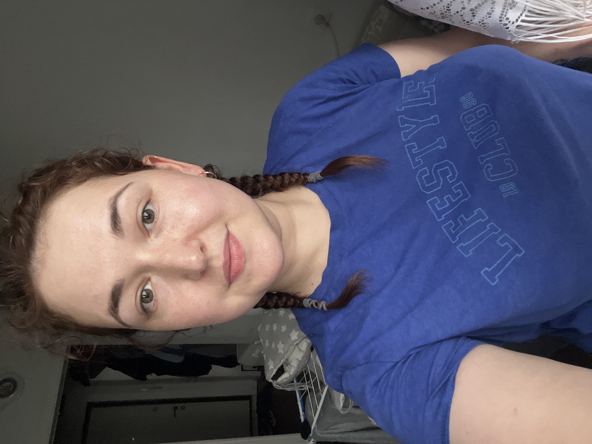 A woman with auburn hair styled in two braids is looking at the camera. She is wearing a blue t-shirt with "LIFESTYLE CLUB 19 96" printed on it. She has a small hoop earring in her left ear. The background shows a dimly lit room with a doorway and clothes hanging on a rack. There is also a white fringed fabric on the right side of the image. The image appears to have been taken indoors.