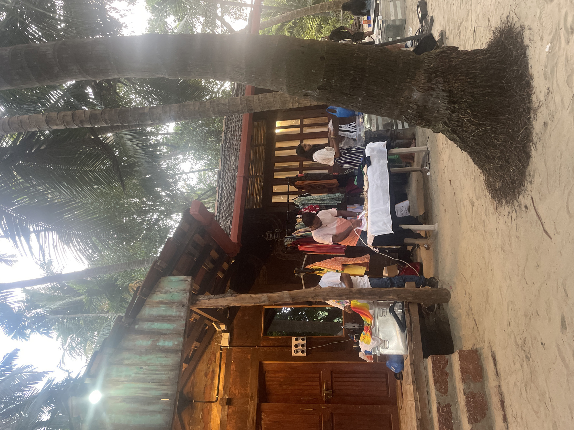 The scene is set in Bhogave, India, near a sandy beach. A wooden structure, possibly a shop, with a tiled roof, is situated under palm trees. People are browsing through clothes displayed on tables outside the shop. Electrical outlets are visible on the shop's wall. Behind the shop, there are trees. The beach sand surrounds the structure.