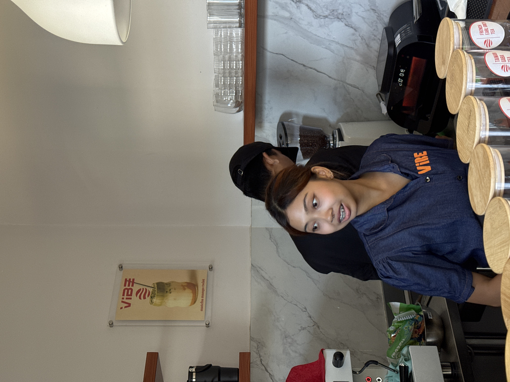 A young woman wearing a blue denim shirt with the word "VIBE" on it, as well as a black hat and a black top, stands at a countertop area in what appears to be a café or shop. She is looking towards the camera. The counter has canisters of teas and a coffee machine to her right. Behind her, there is a coffee grinder, water glasses mounted on a wall, and a menu poster that says "VIBE matcha mango twist" displayed on the wall. There is some equipment at the bottom of the image, along with a red towel. The scene is located in Denpasar, Indonesia.