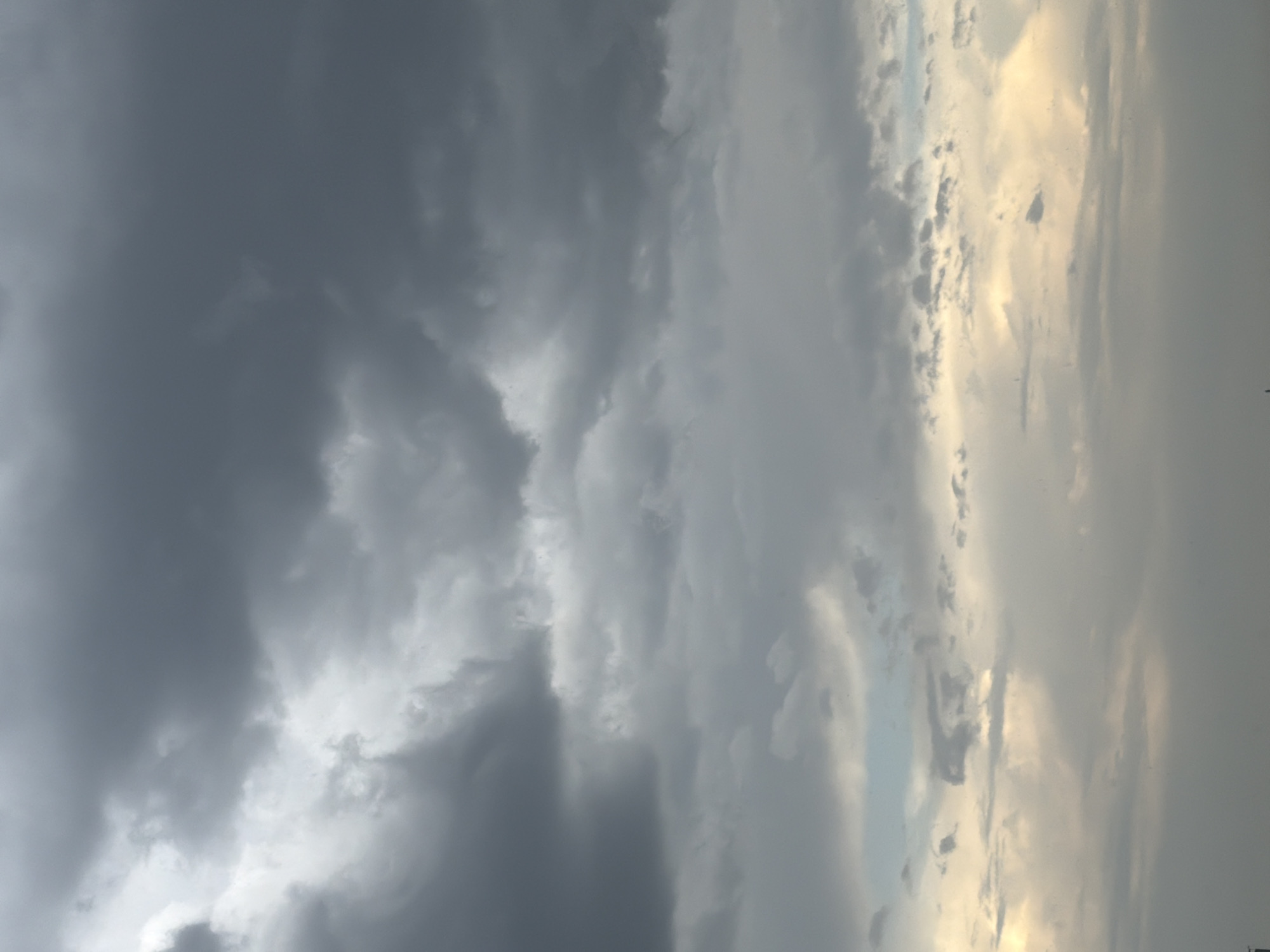 The image captures a dramatic sky filled with a mixture of dark, heavy storm clouds and lighter, more wispy formations. The clouds are varied in texture and density, creating a sense of depth and movement. The lower portion of the sky shows a brighter, yellowish hue, suggesting the presence of sunlight breaking through or reflecting off the clouds. There is no visible indication of the city of Pavlodar, Kazakhstan, or any specific landmarks. No people, subjects, or actions are discernible within the frame, making it a purely atmospheric landscape.